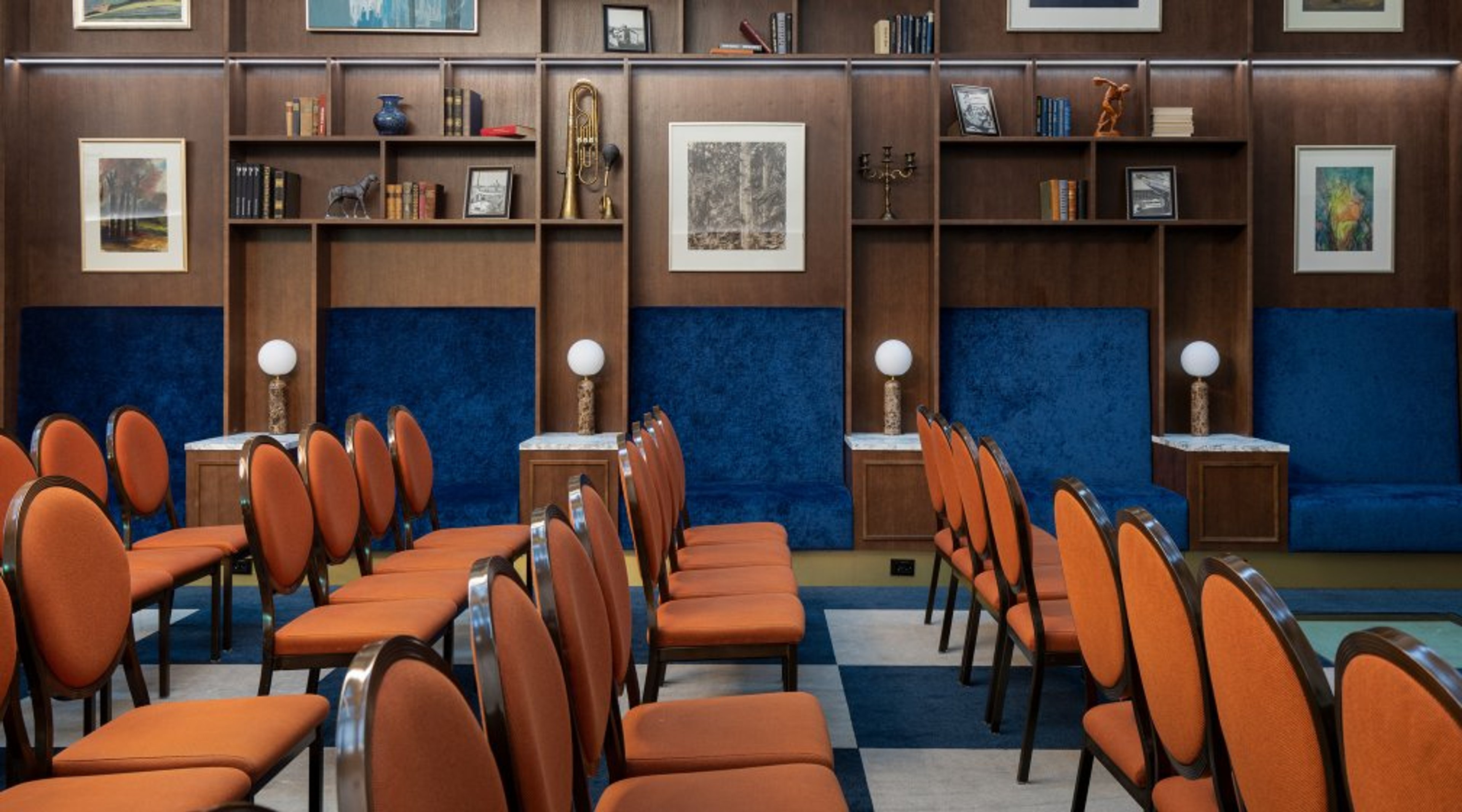 Kokoushuone, jossa on oranssiverhoiltuja tuoleja, siniset seinäpaneelit ja takaseinällä puinen kirjahylly koristeineen ja taideteoksineen.