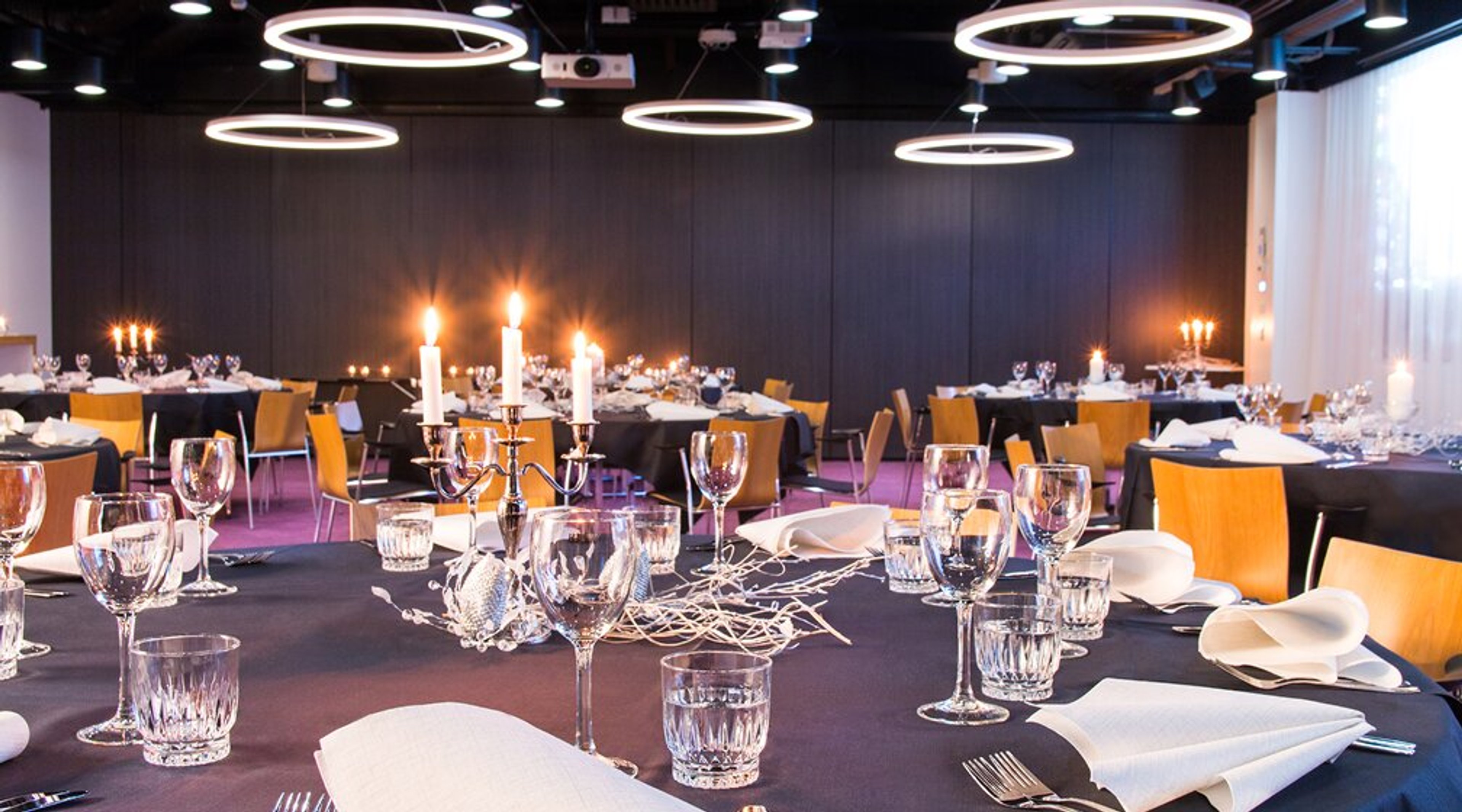 Peoruum laudadega, küünlajalad ja lauanõud. Mustad laudlinad, puidust toolid ja rõngakujulised laevalgustid.