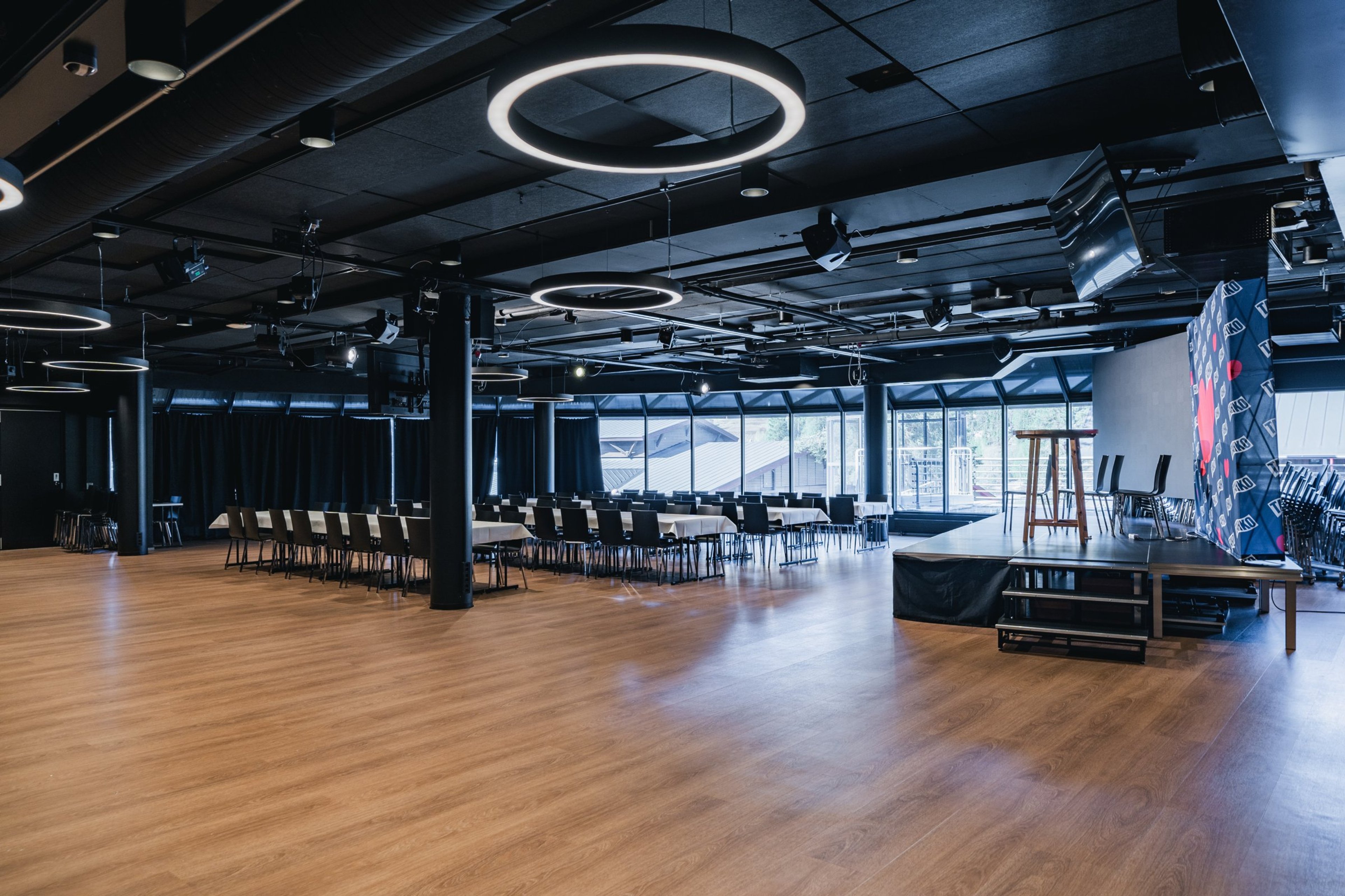 Moderni tapahtuma- tai konferenssitila jossa puulattia, mustia pöytiä, rivi-istuinjärjestys ja lava. Korkea musta katto rengasmaisilla valoilla.