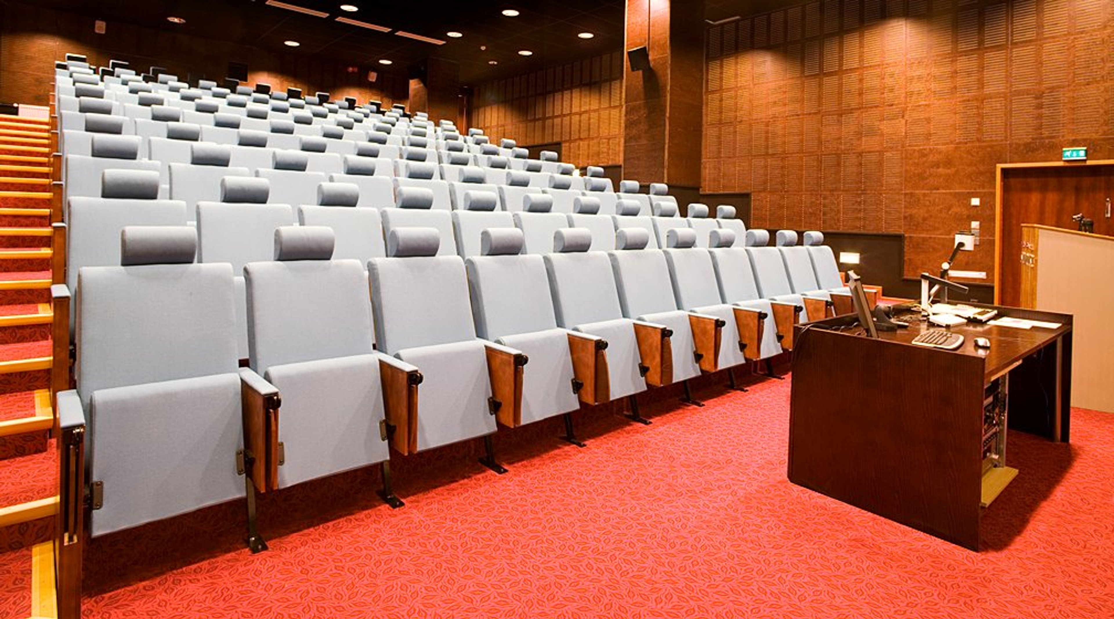 Auditorio, jossa harmaat pehmustetut istuimet nousevilla riveillä, puupaneeliseinät ja punainen kokolattiamatto. Edessä puhujanpöytä.