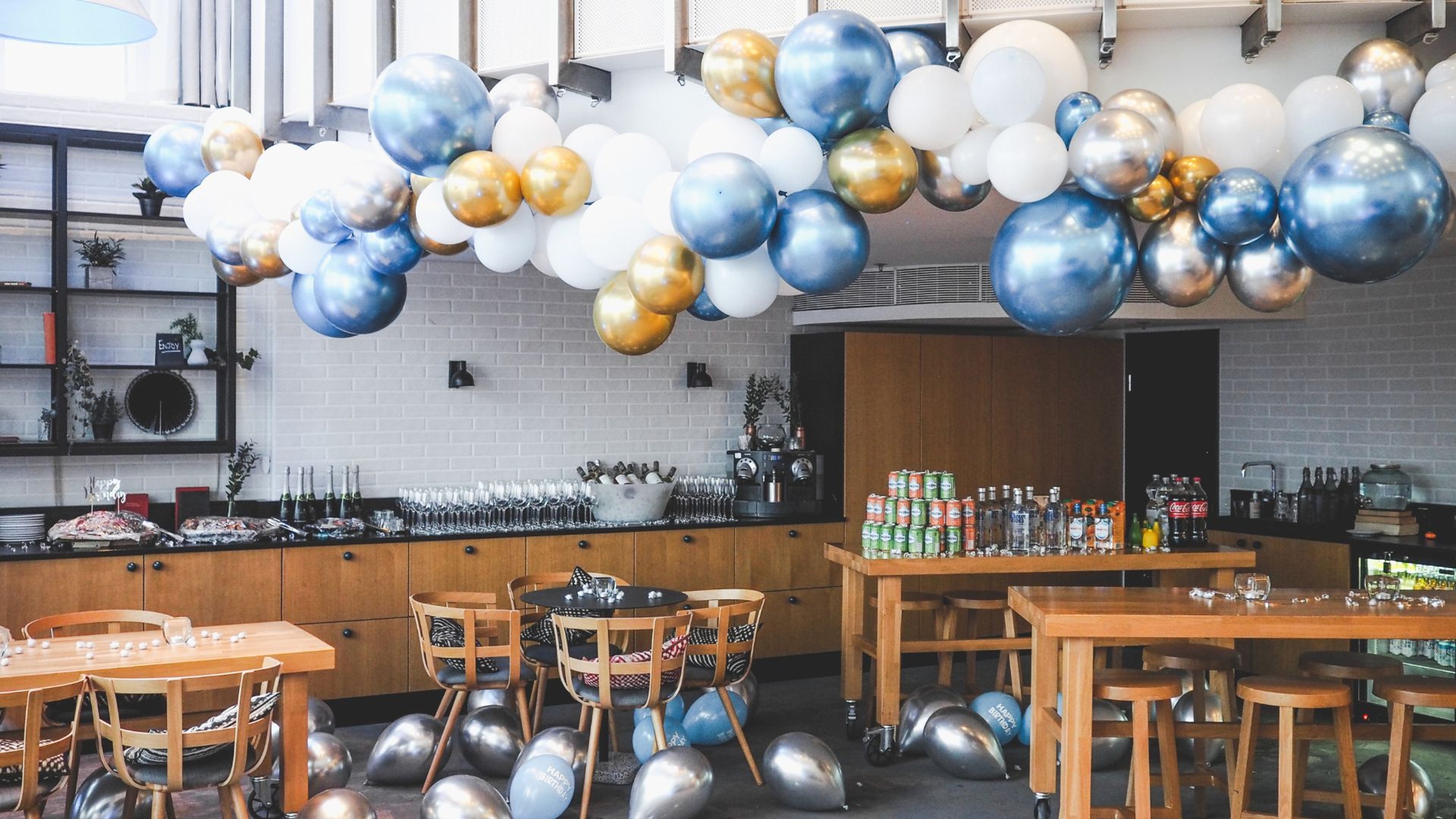 Juhlatila koristellu sinisillä, kultaisilla ja valkoisilla ilmapalloilla. Puisia pöytiä ja tuoleja, tiskillä juomia ja laseja.