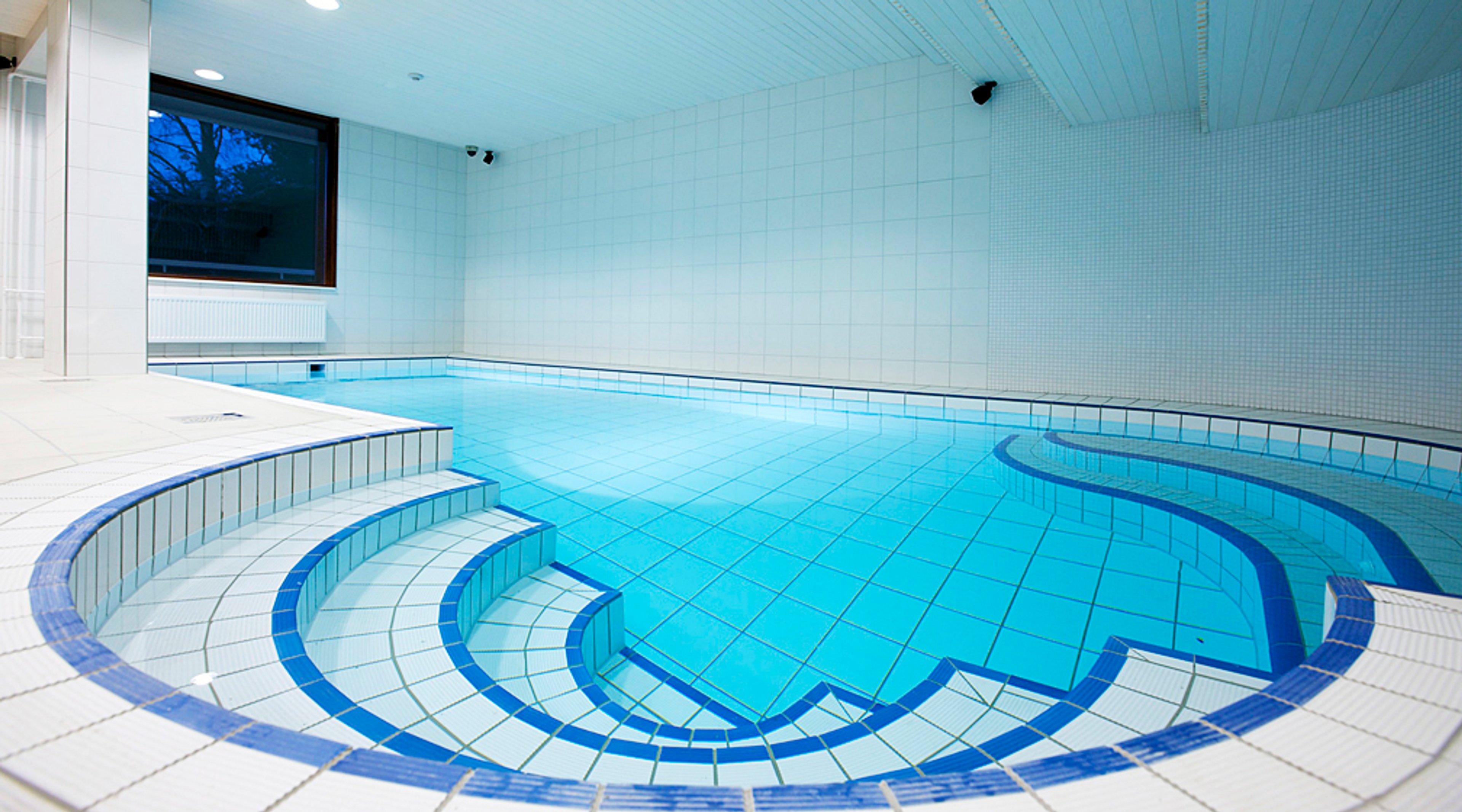 Sisäuima-allas valkoisin ja sinisin laatoitetulla alueella. Altaaseen johtaa portaat ja ison ikkunan kautta näkyy ulos.