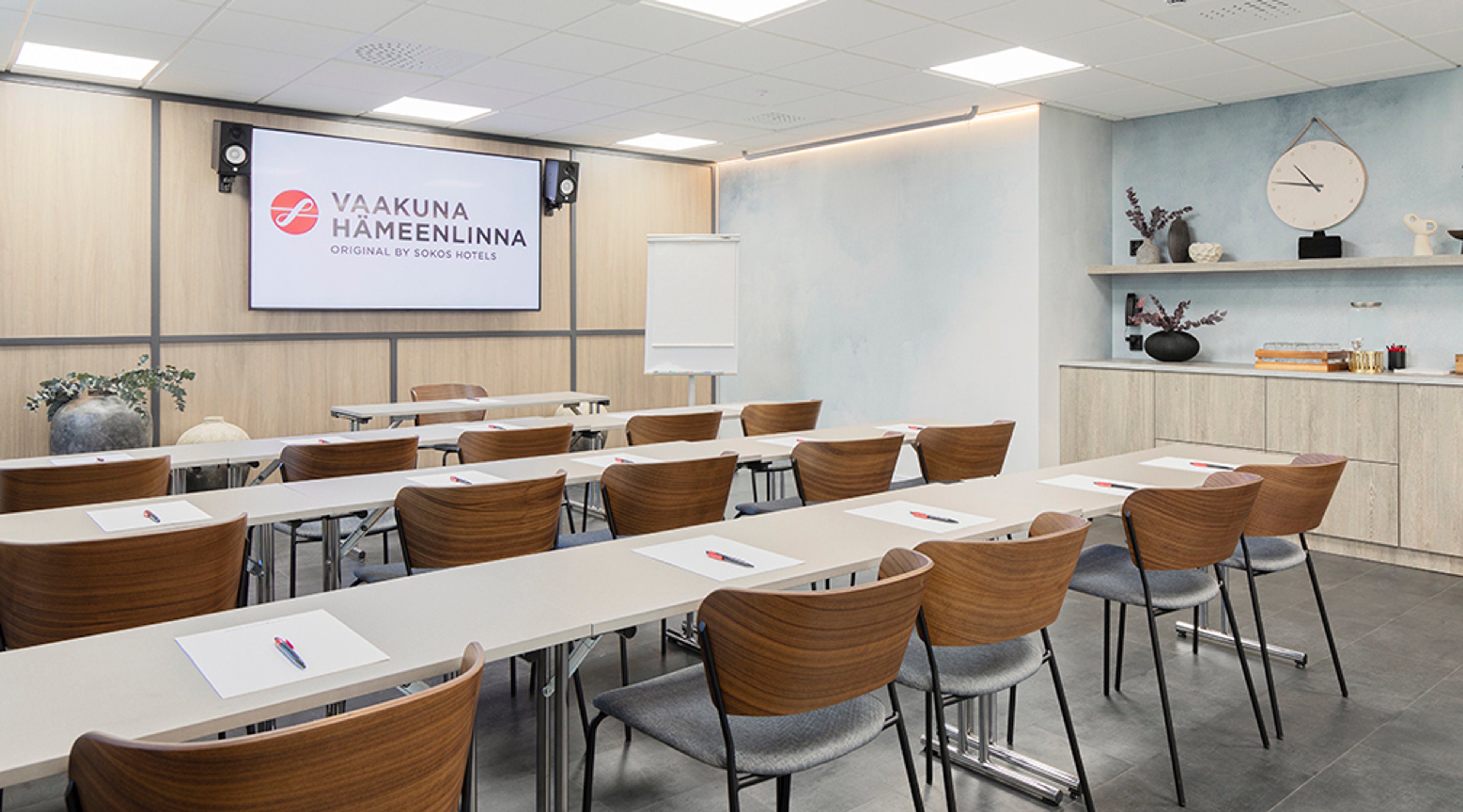 Moderni kokoustila vaaleilla pöydillä ja puisilla tuoleilla. Edessä suuri näyttö, takaosassa pieni hylly ja seinäkello.