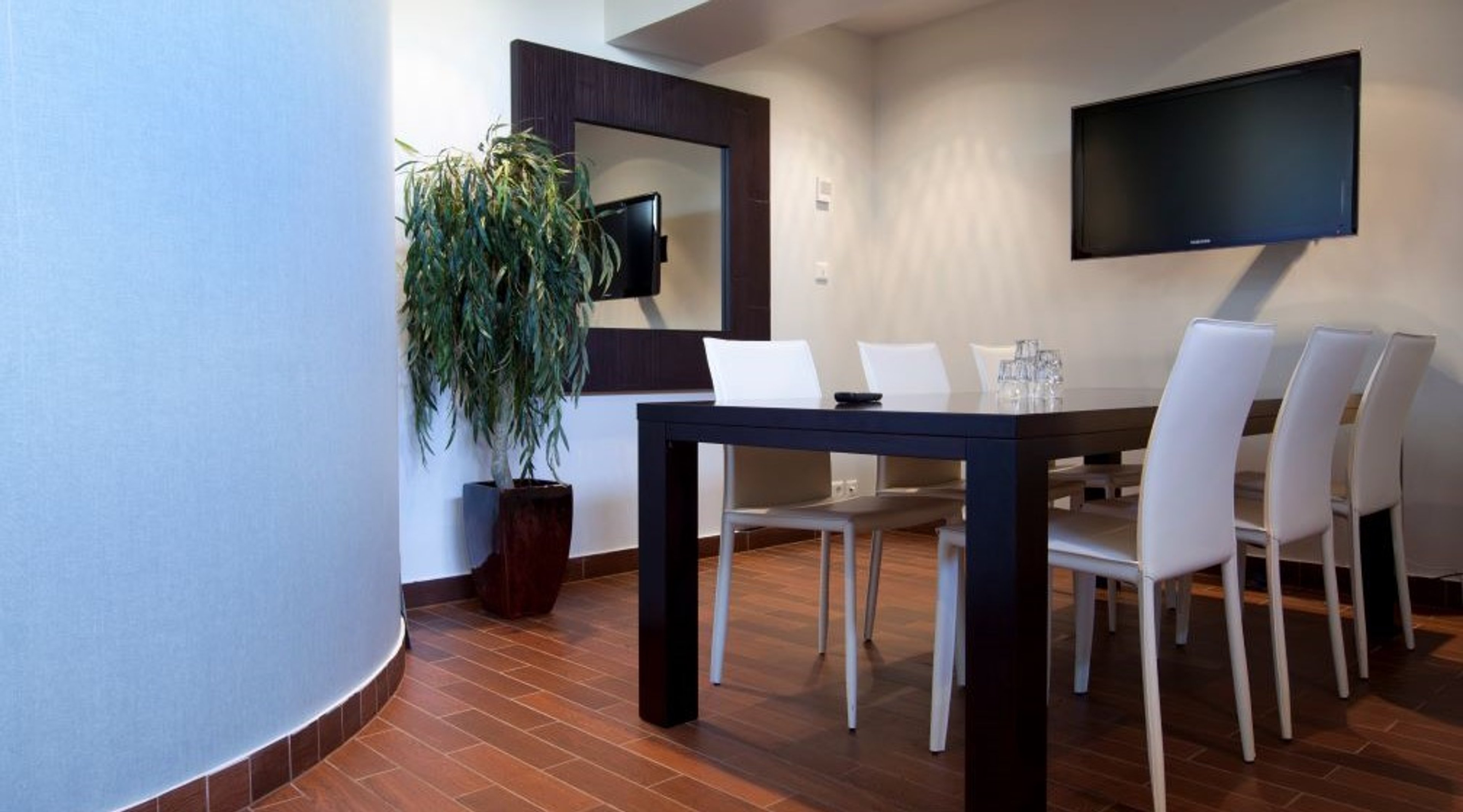 Moderni neuvotteluhuone, jossa tumma pöytä, valkoiset tuolit, lattia-kasvi, tv-näyttö seinällä ja puulattia.