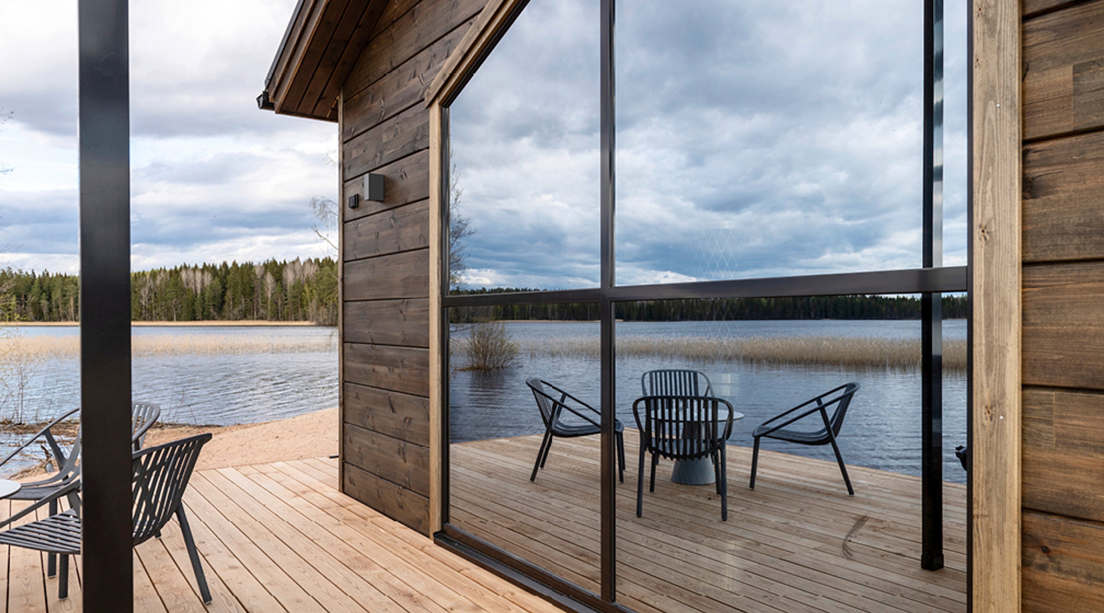 Puinen laituri järven rannalla, jossa mustia tuoleja. Suurista ikkunoista näkyy järvimaisema ja pilvinen taivas metsän yllä.