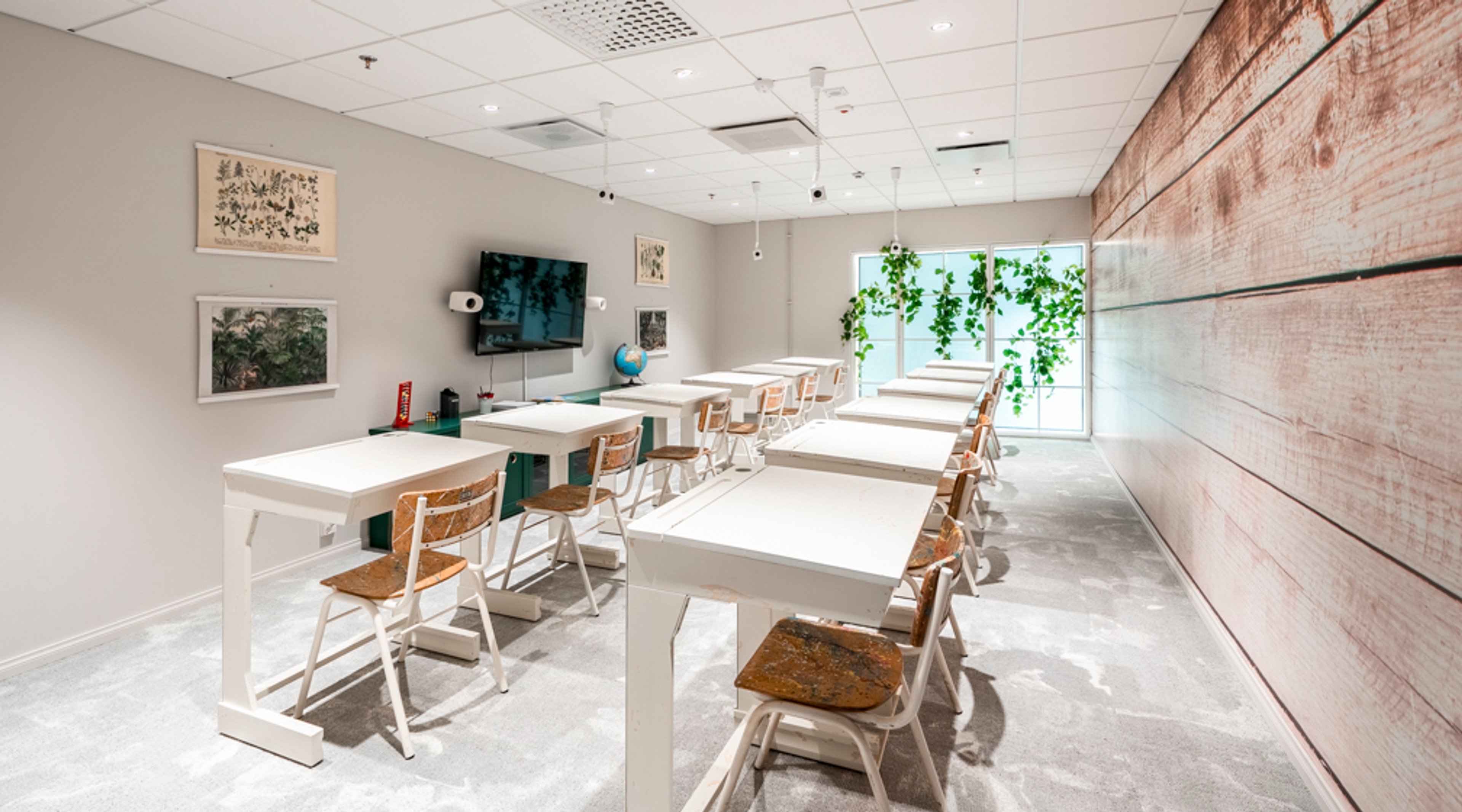 Moderni kokoustila, jossa valkoiset pöydät ja puiset tuolit. Seinällä on televisio ja ikkunan äärellä viherkasveja. Tilassa kova lattia ja hyvä valaistus.