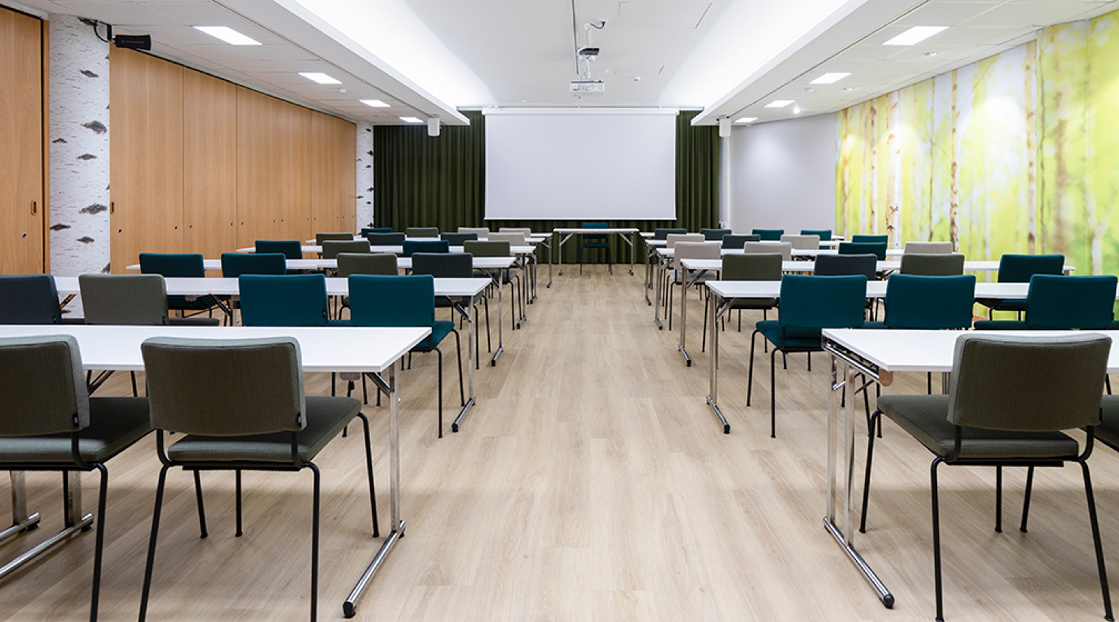 Valoisa kokoushuone jossa vaaleat pöydät, siniset ja harmaat tuolit, valkoinen esityskangas ja puulattia. Seinillä puupaneeli ja metsäaiheinen kuva.