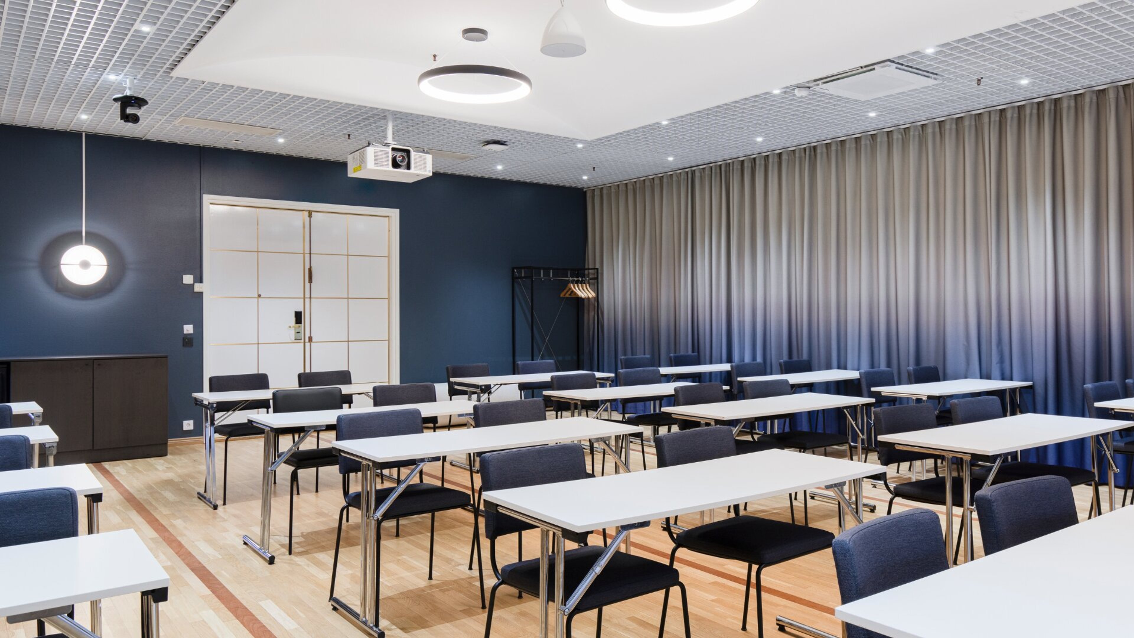 Modern meeting room with blue walls, white tables and chairs. Wooden floor, good lighting and ceiling-mounted projector system.