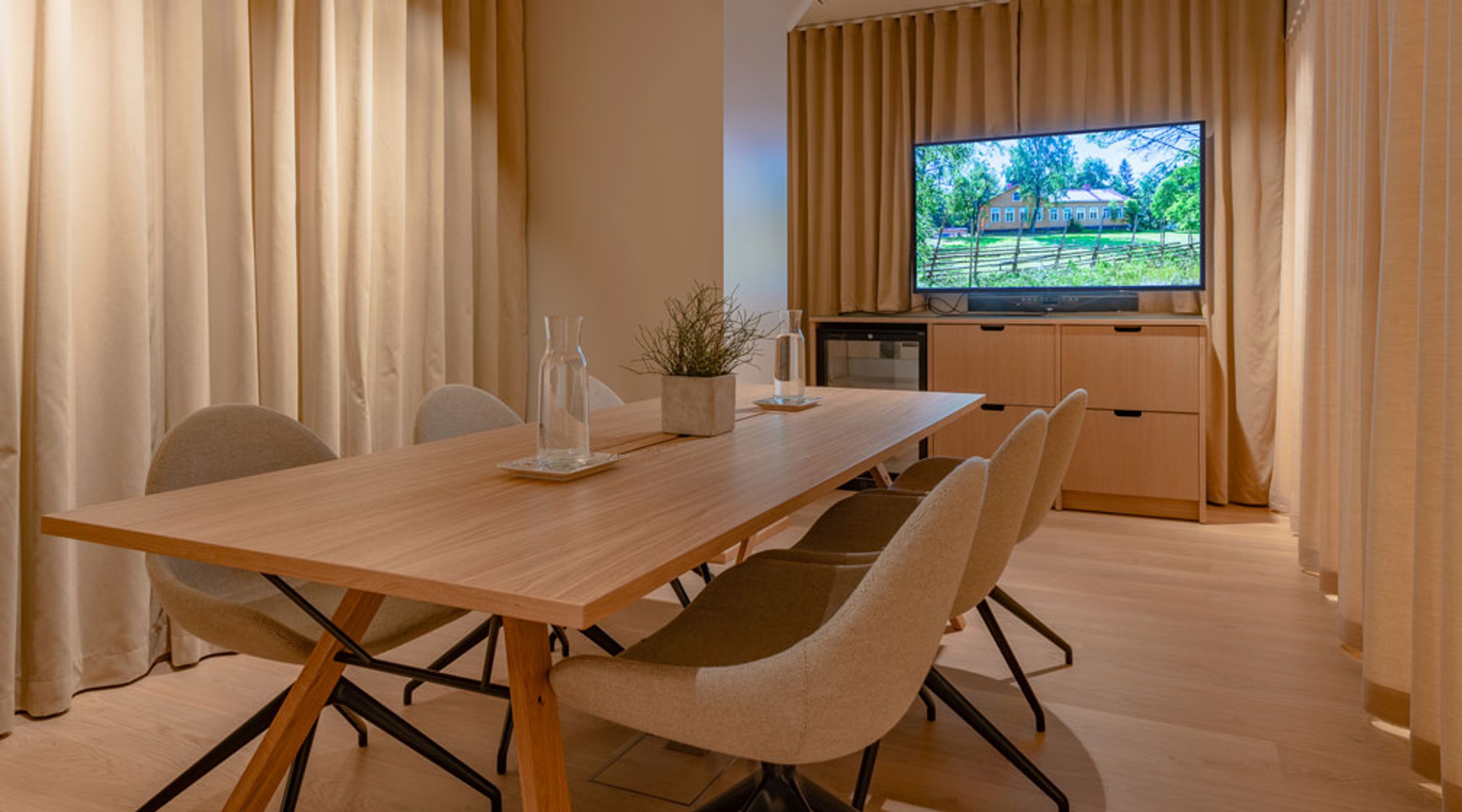Moderni neuvotteluhuone puisella pöydällä ja pehmeillä tuoleilla. Taustalla televisio ja beigen sävyiset verhot. Vaalea puulattia.