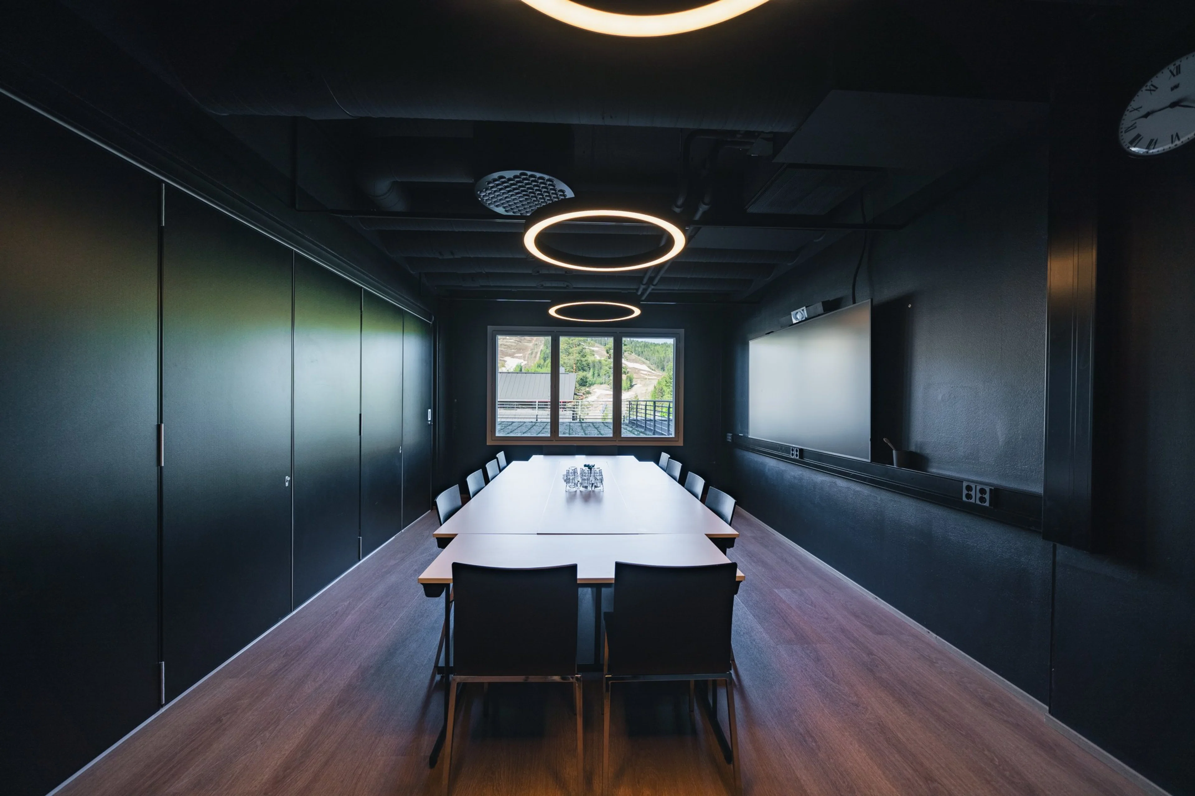 Tummasävyinen neuvotteluhuone, jossa pitkä puinen pöytä, tuoleja, näyttö seinällä ja pyöreät valot katossa. Ikkunasta näkyy maisema.