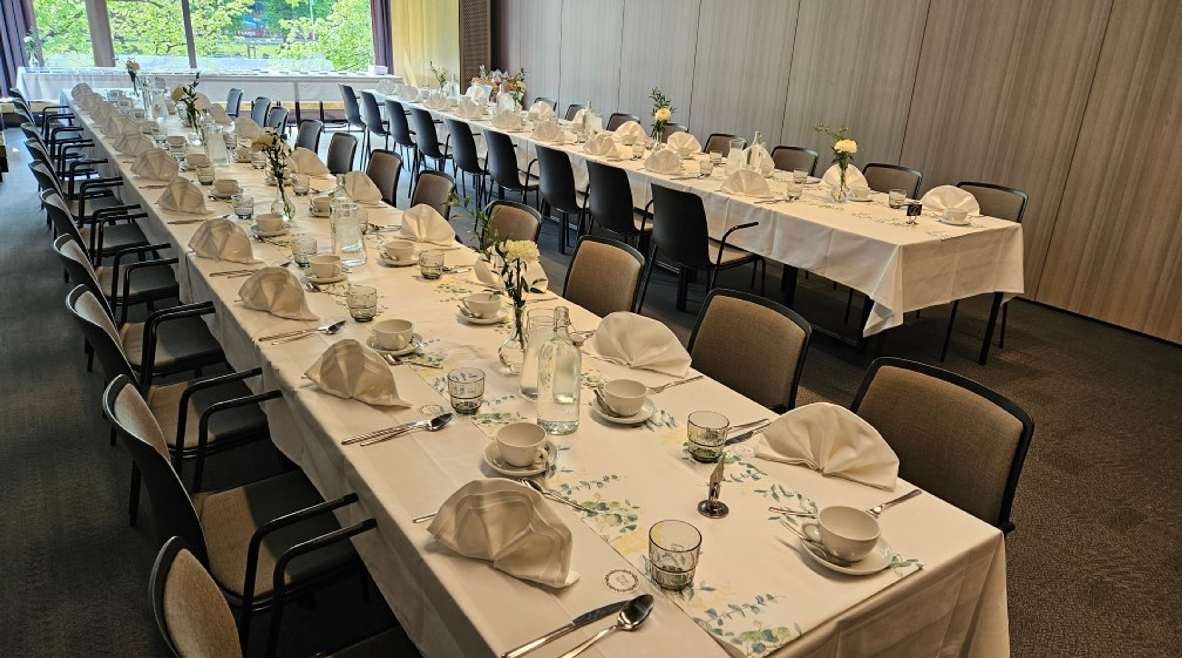 Juhlavasti katetut pitkät pöydät ruokasalissa. Pöydillä on valkoiset liinat, taitetut servietit ja pieniä kukka-asetelmia. Vieressä suuret ikkunat.