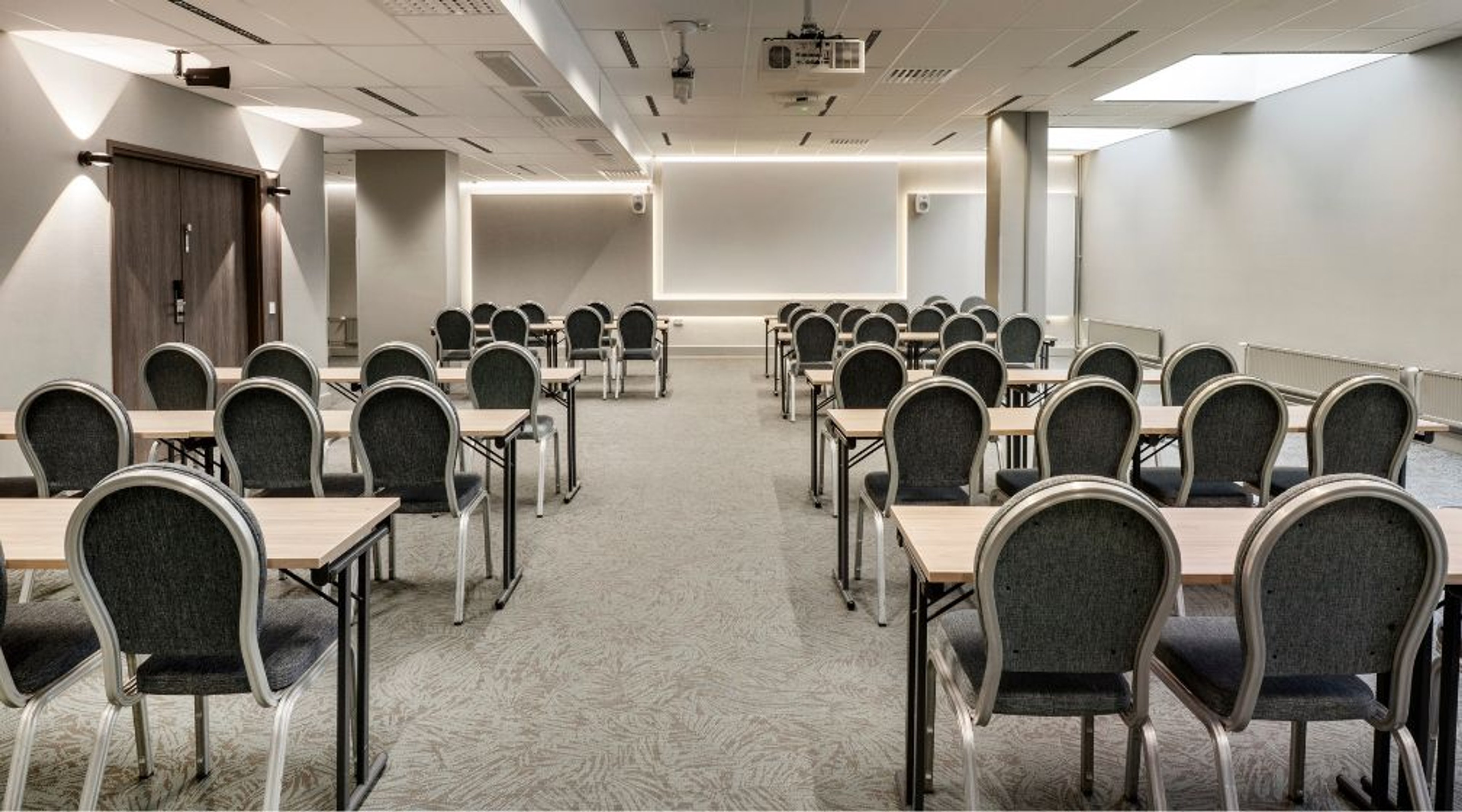 Kokoustila, jossa on rivimuodostelmassa pöytiä ja tuoleja. Huoneessa on tasainen lattia, hyvä valaistus ja puinen sisäänkäyntiovi.