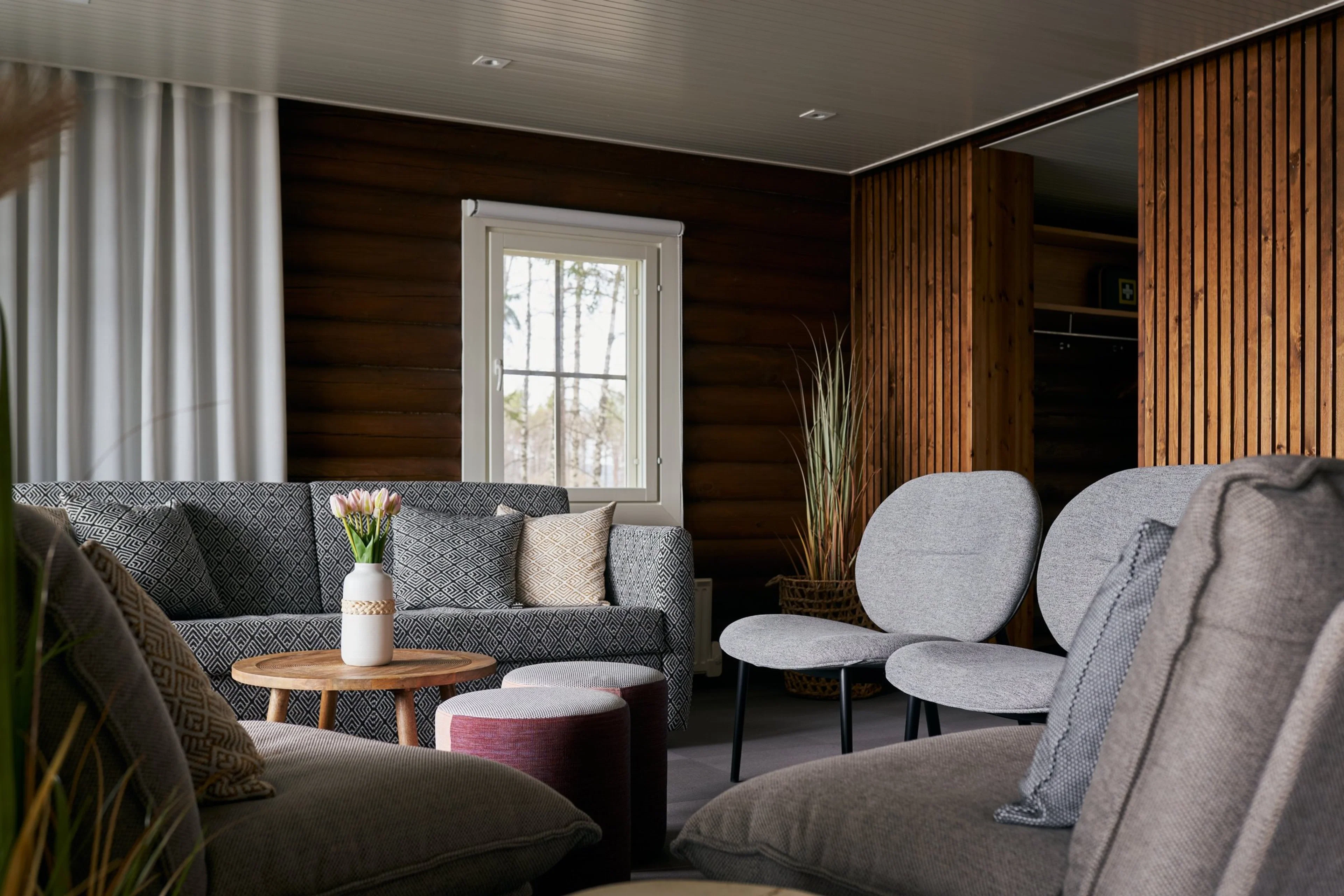 Kotoisa olohuone puuseinillä, harmaa sohva kuviollisilla tyynyillä, pyöreä puupöytä maljakolla, kaksi harmaata tuolia ja ikkunasta näkyy metsä.