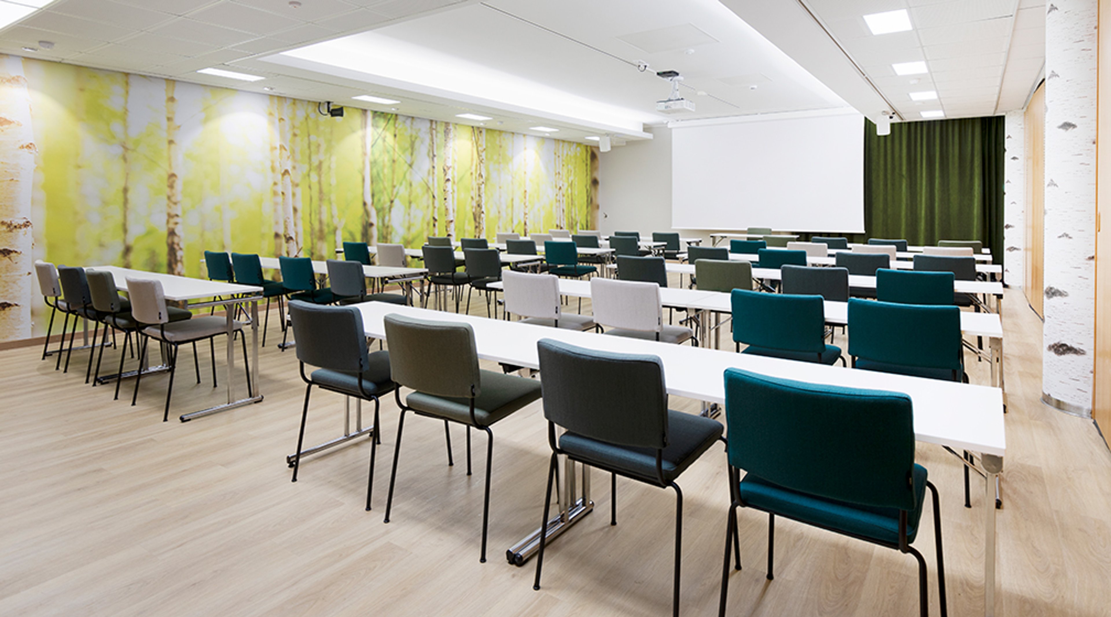 Kokoushuone pitkillä pöydillä ja erivärisillä tuoleilla. Seinällä koivumetsäaiheinen taustatapetti. Tasainen puulattia ja valkoiset pöydät.