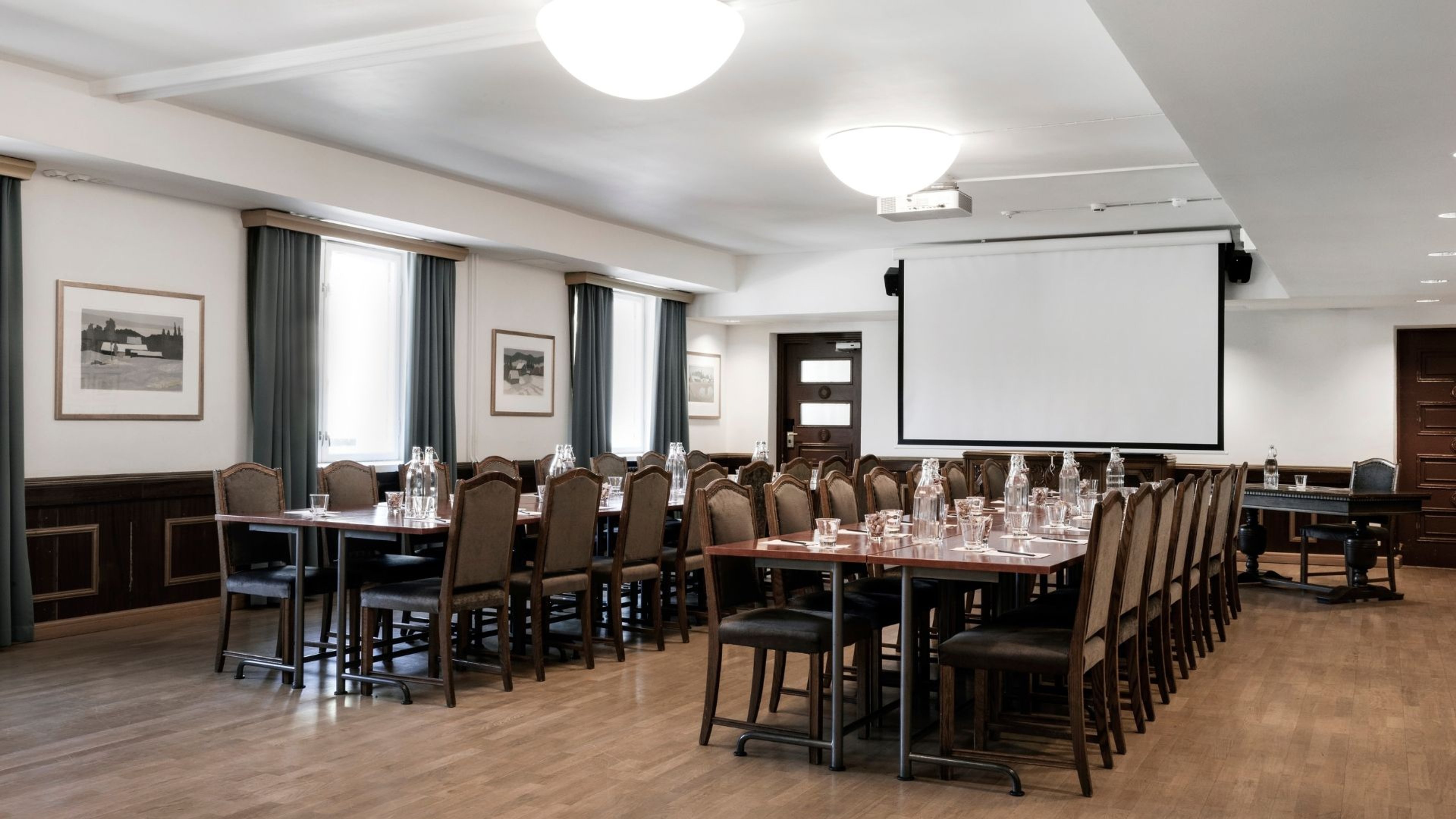 Kokoushuone, jossa on pöytiä tuoleineen, projektori valkokankaalla ja puulattia. Tilassa on suuret ikkunat tummilla verhoilla.