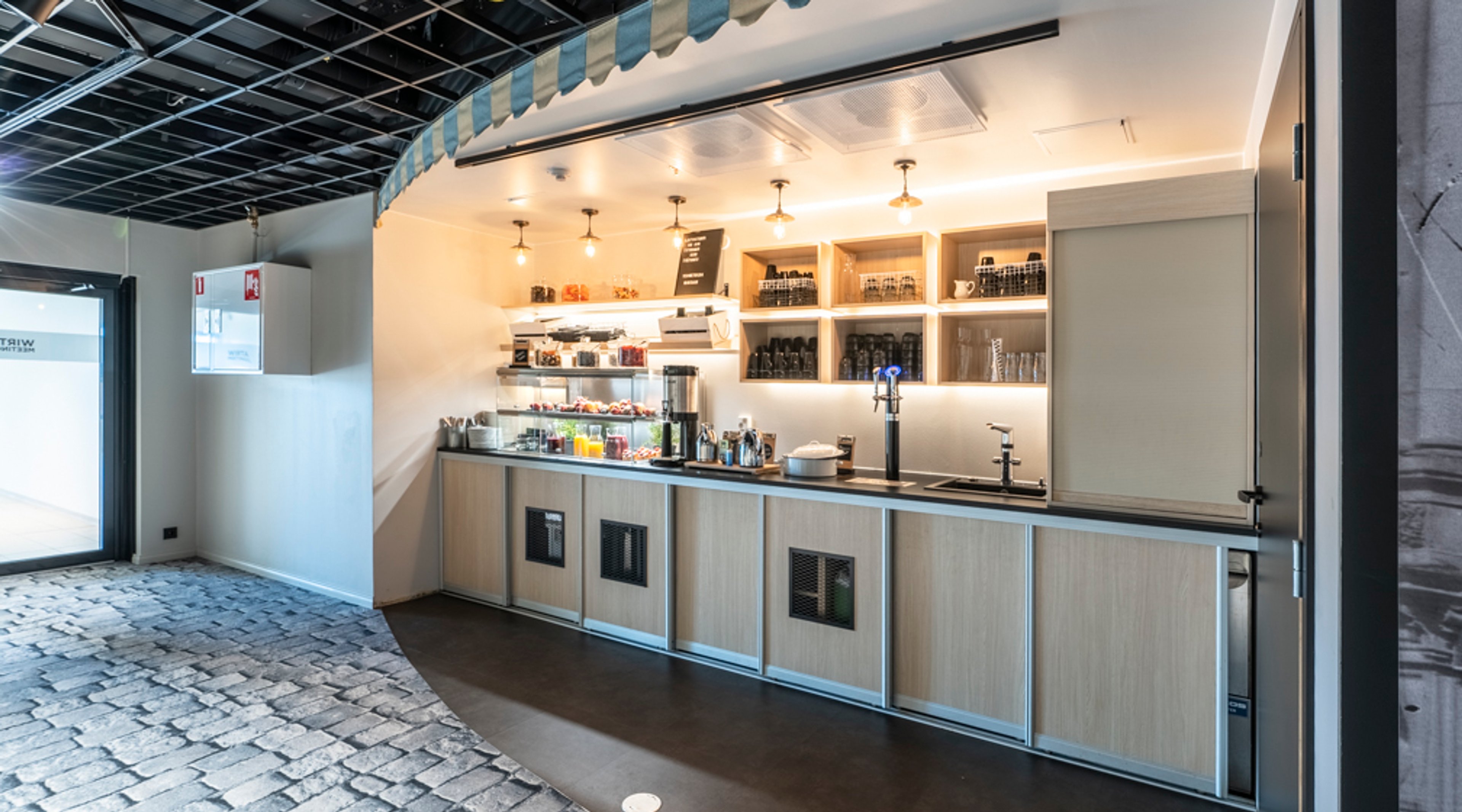 Kahvilatiski puisilla kaapeilla, taustalla lasihyllyjä. Lattia kivikuviollinen, huone on avara ja hyvin valaistu.