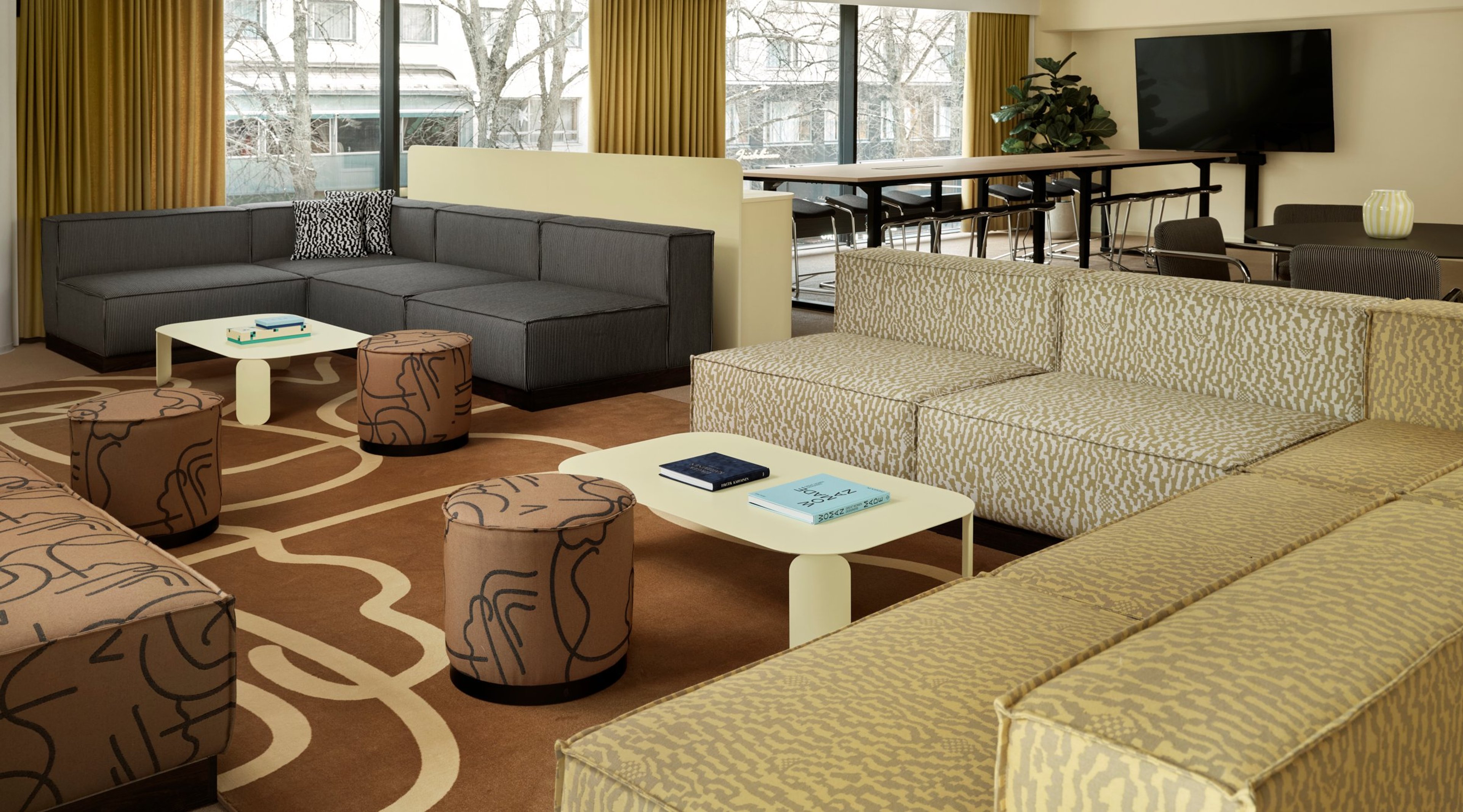 Moderni oleskelutila, jossa harmaa ja kuviollinen beige sohva, pyöreitä raheja ja matalat pöydät. Taustalla neuvottelupöytä ja televisio. Isot ikkunat ja tasainen lattia.