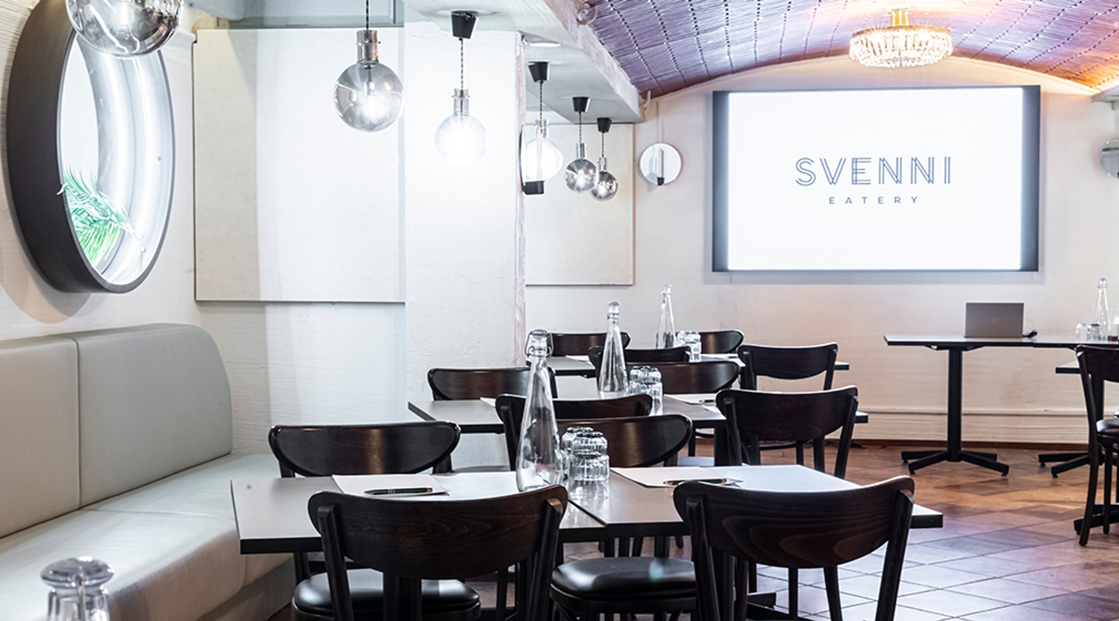 Moderni ravintolan sisätila valkeilla seinillä, mustilla tuoleilla ja tummalla laattalattialla. Kattokruunu ja erilaiset valaisimet luovat tunnelmaa.