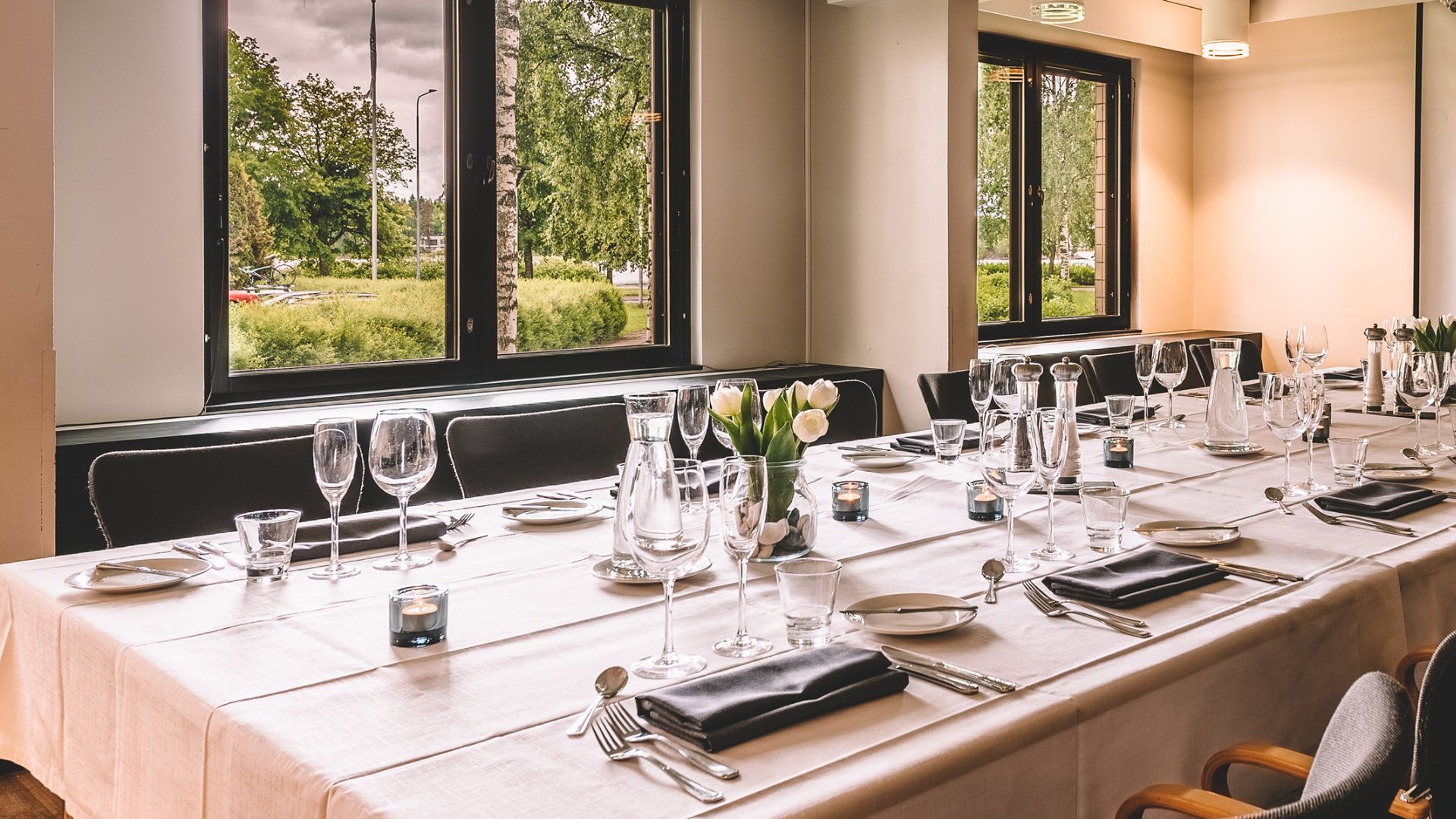 Elegantisti katettu pöytä ravintolassa, jossa suurista ikkunoista avautuu näkymä vehreälle piha-alueelle.