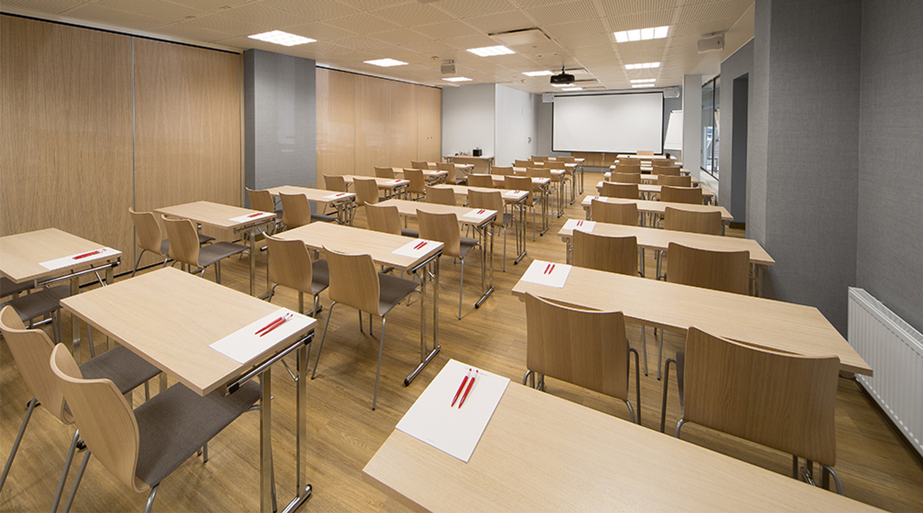 Moderni seminaarihuone puisilla pöydillä ja tuoleilla, valkoinen projektoriruutu edessä ja tasainen puulattia.