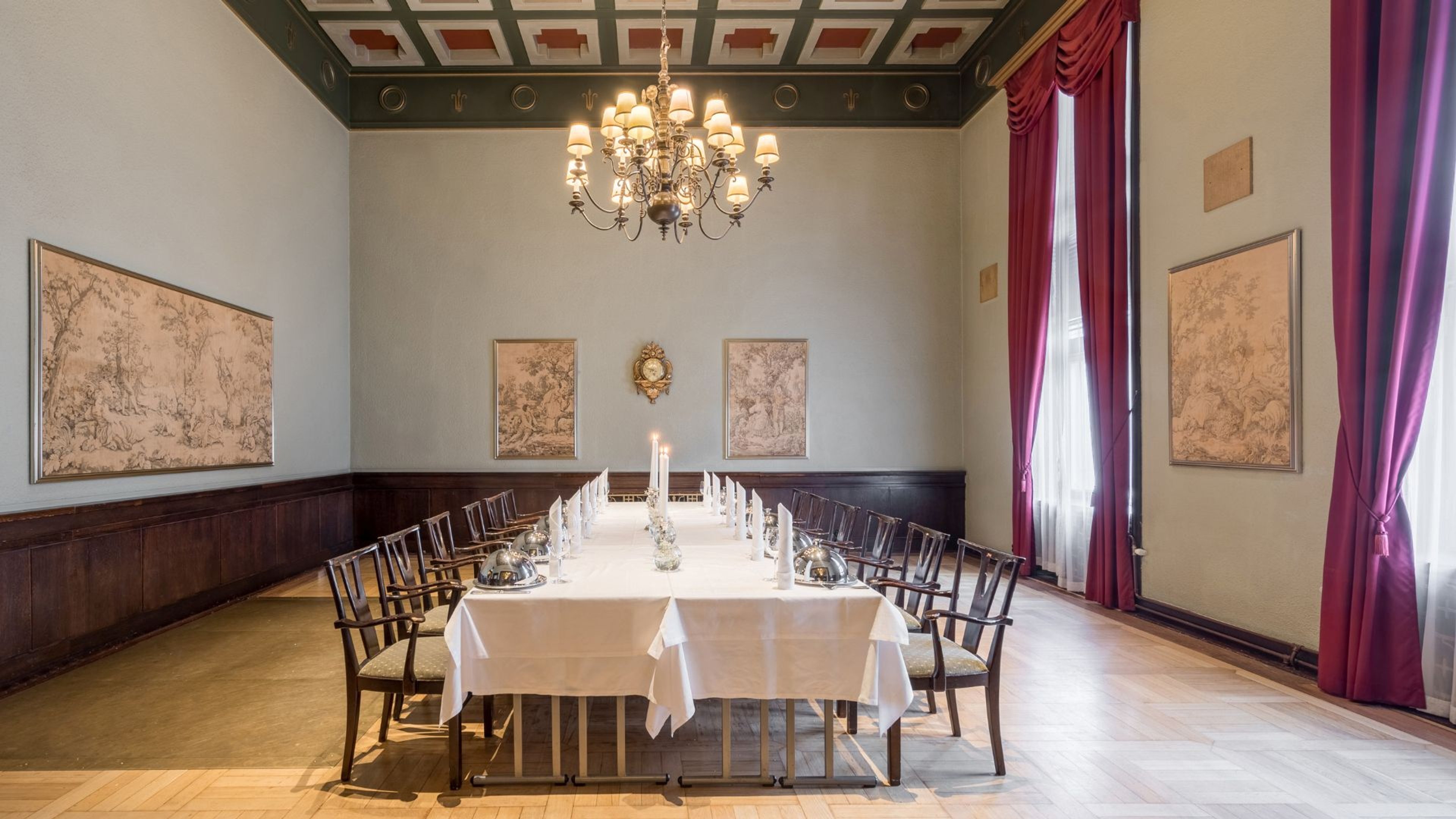 Elegantti juhlahuone, jossa pitkä pöytä valkoisella liinalla ja tuoleja. Koristeellinen kattokruunu, punaiset verhot ja taidepiirroksia seinillä.