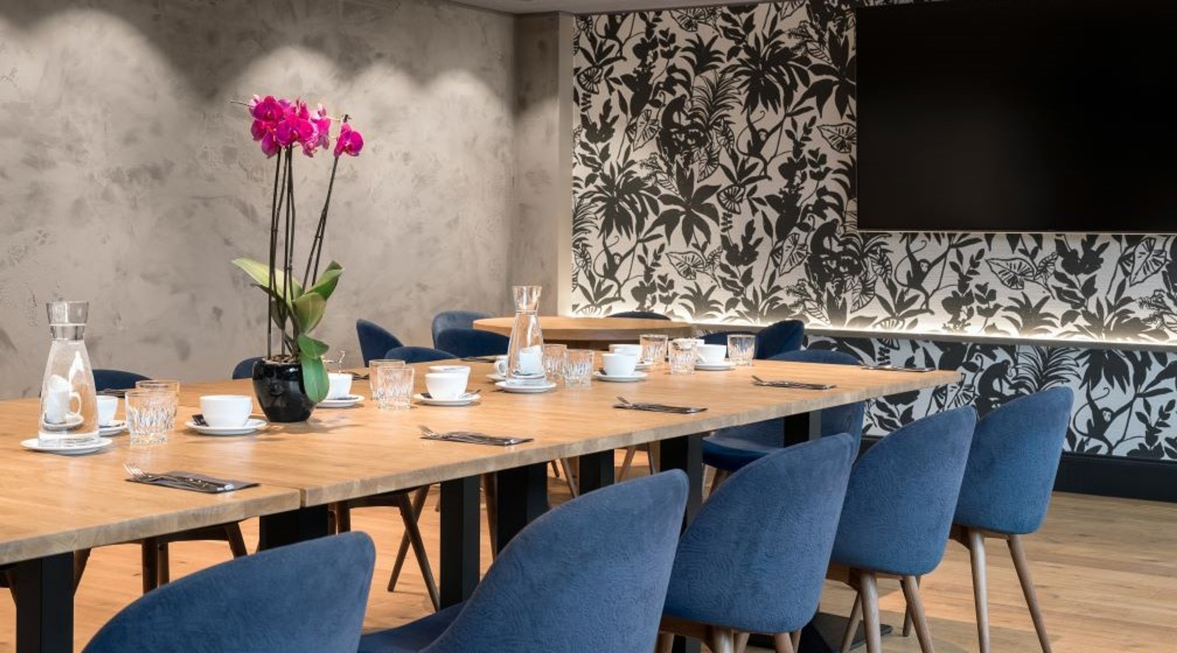 Kokoushuone, jossa pitkä puupöytä ja sinisiä tuoleja. Pöydällä on laseja, kahvikuppeja ja pinkki orkidea. Takaseinässä on musta-valkoinen kukkatapetti.