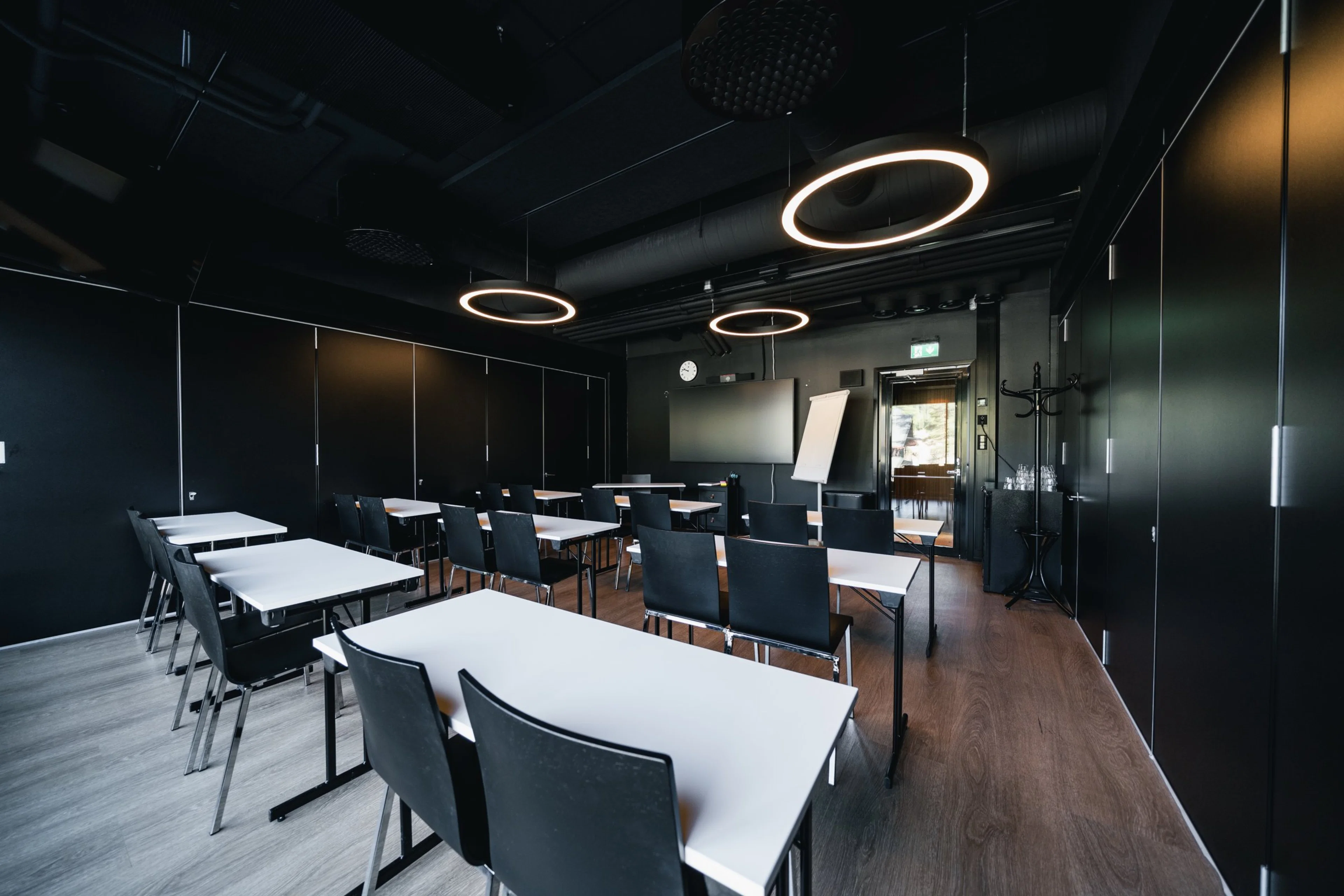Moderni kokoustila mustilla seinillä, valkoisilla pöydillä ja rengasmaisilla kattovalaisimilla. Tilassa on puulattia ja näyttö edessä.