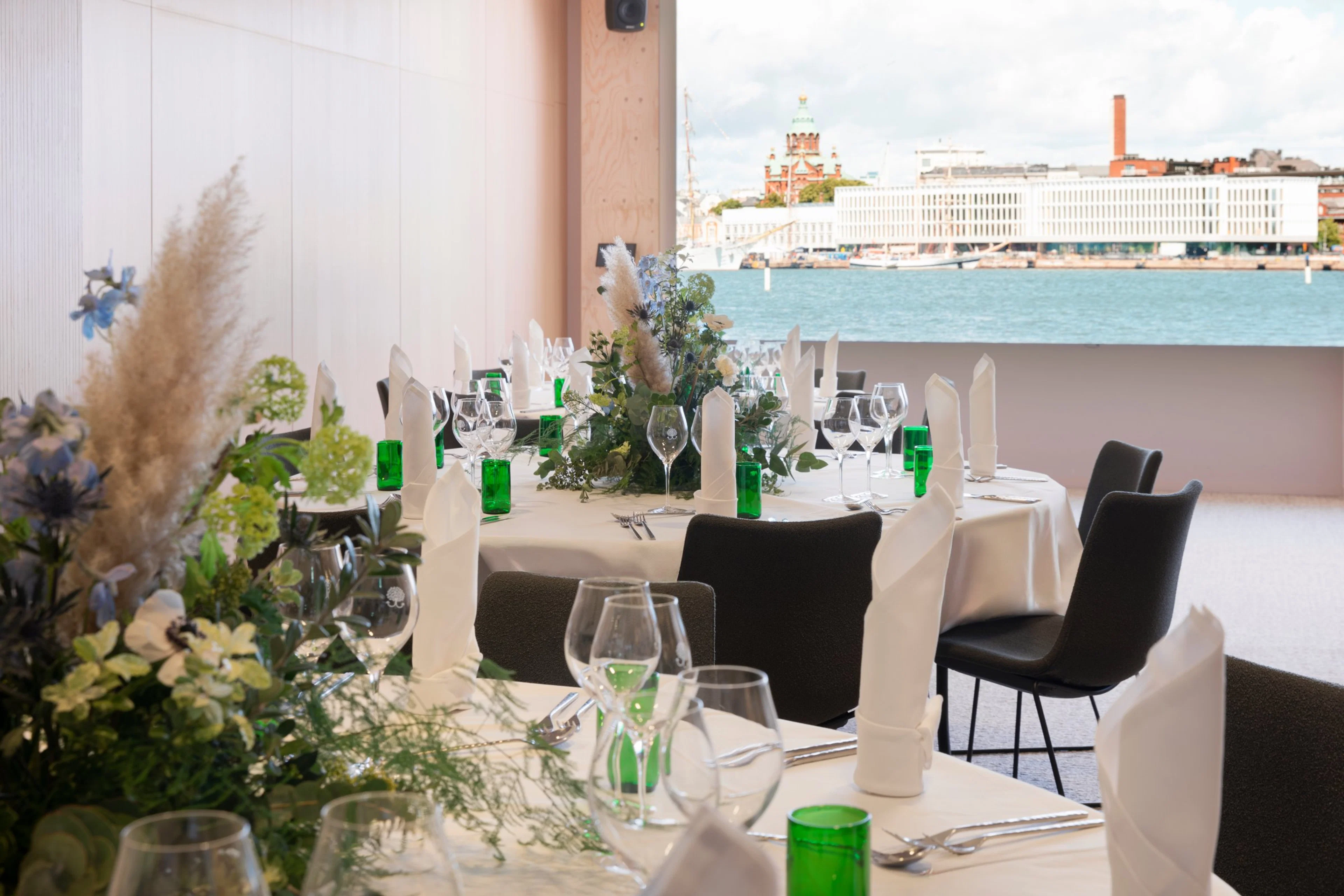 Elegant restaurang med havsutsikt. Bord dukade med vita dukar, gröna glasflaskor och blomsterarrangemang. Uspenskijkatedralen i Helsingfors syns genom fönstret.