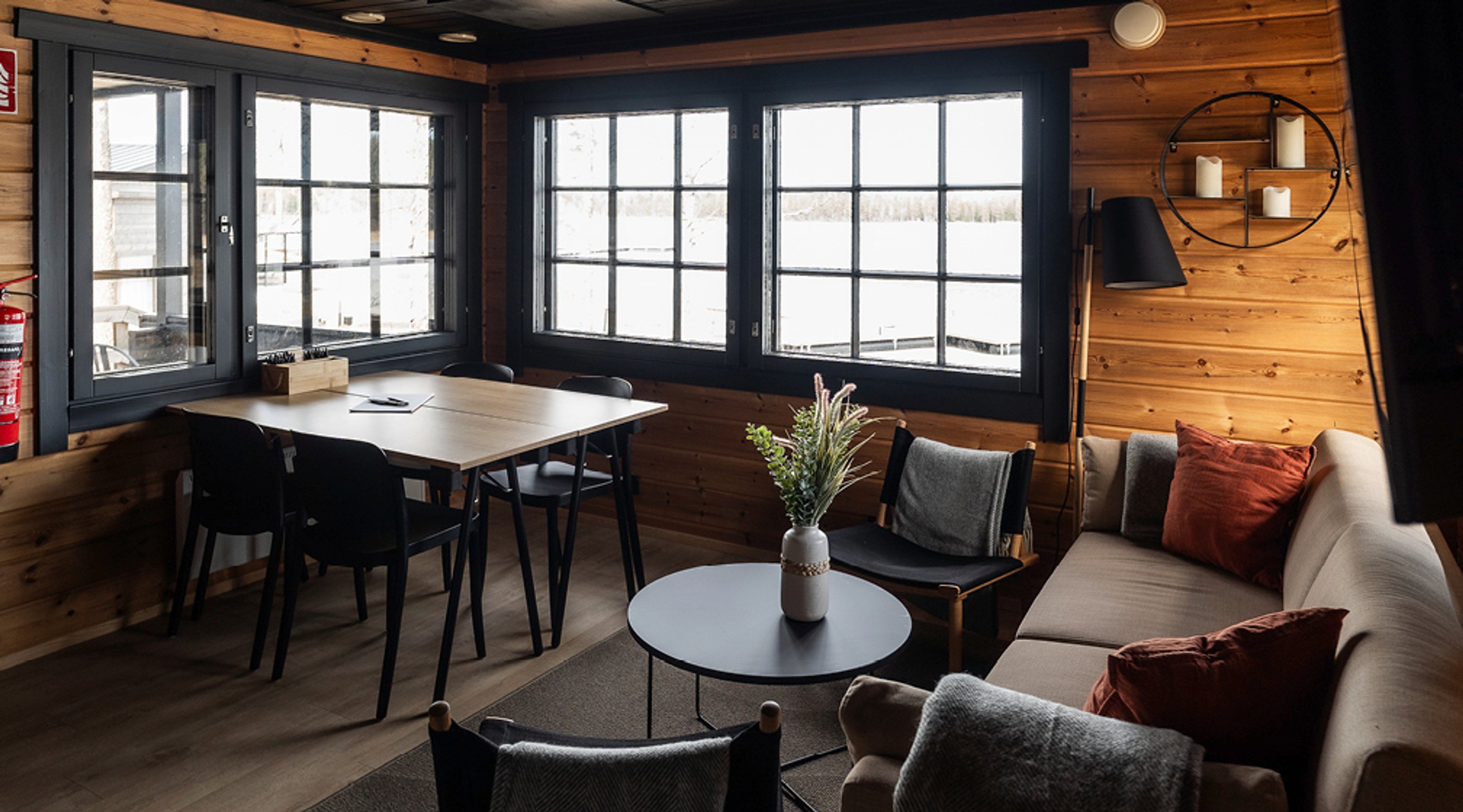 Valoisa kokoustila puuseinillä, jossa sohvat, pyöreä pöytä ja ruokapöytä tuoleineen. Isot ikkunat tarjoavat näkymän ulos.