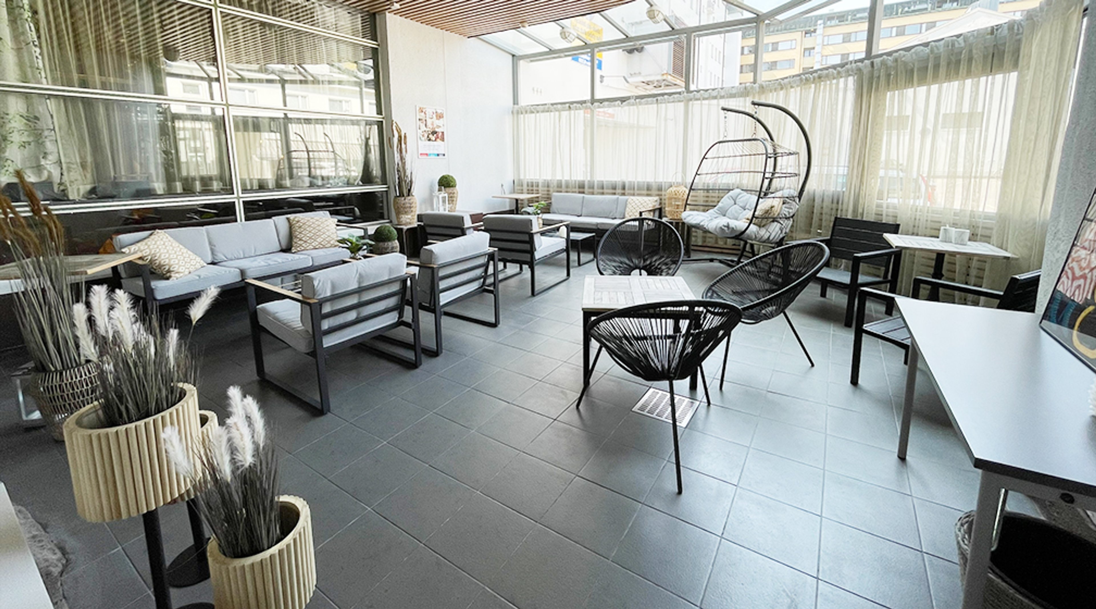 Moderni lasitettu terassi, jossa on tyylikkäitä huonekaluja: vaaleita sohvia, mustia rottinkituoleja ja riippukeinu. Lattia on tummaa laattaa.