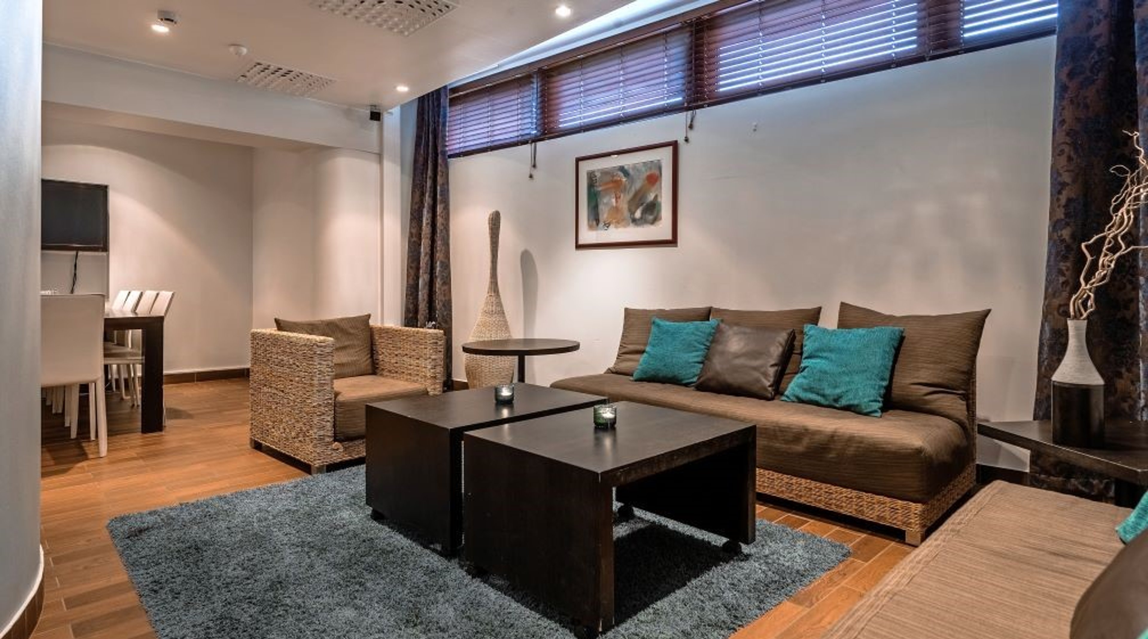 Moderni oleskelutila rottinkihuonekaluilla, ruskealla sohvalla ja turkooseilla tyynyillä. Taustalla näkyy ruokailutila ja puulattia.