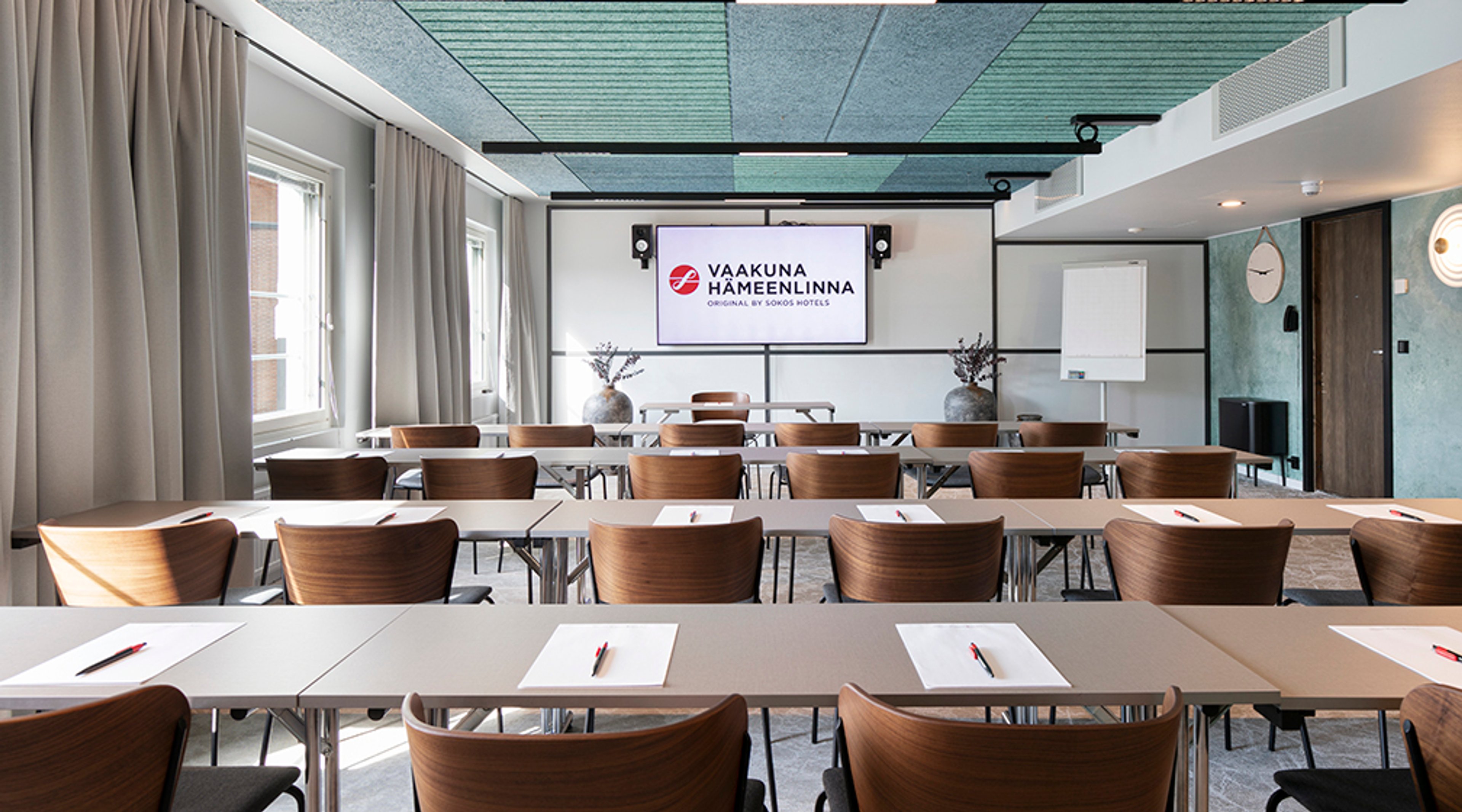 Moderni kokoushuone puisilla tuoleilla, valkoisilla pöydillä ja suurella näytöllä. Tilassa sinivihreä kattopaneeli ja luonnonvaloa ikkunoista.