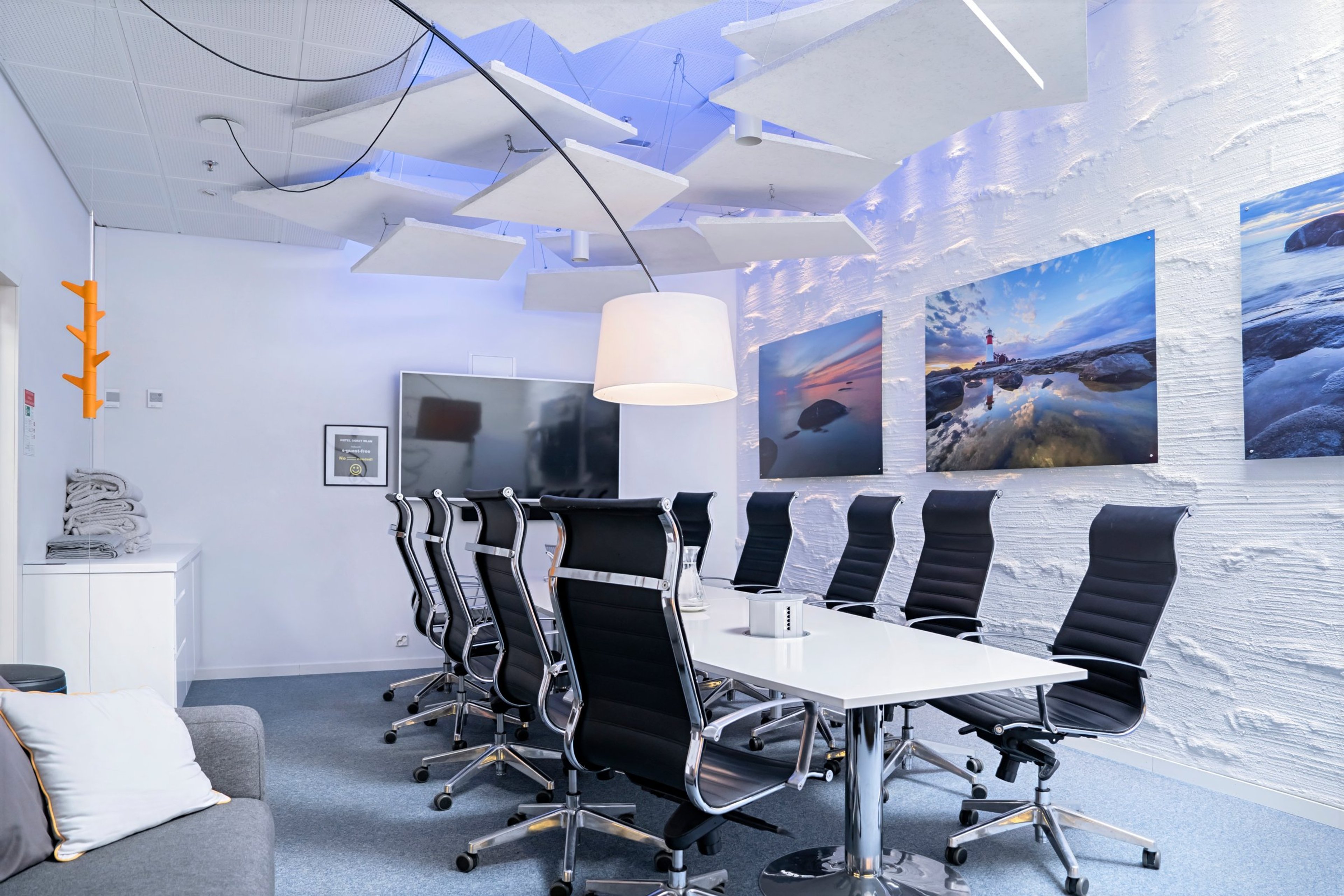 Modernisti sisustettu valkoinen kokoushuone, jossa pitkä pöytä, mustat tuolit ja seinällä merimaisemakuvia. Katosta riippuu valkoinen lamppu ja akustiikkalevyjä.