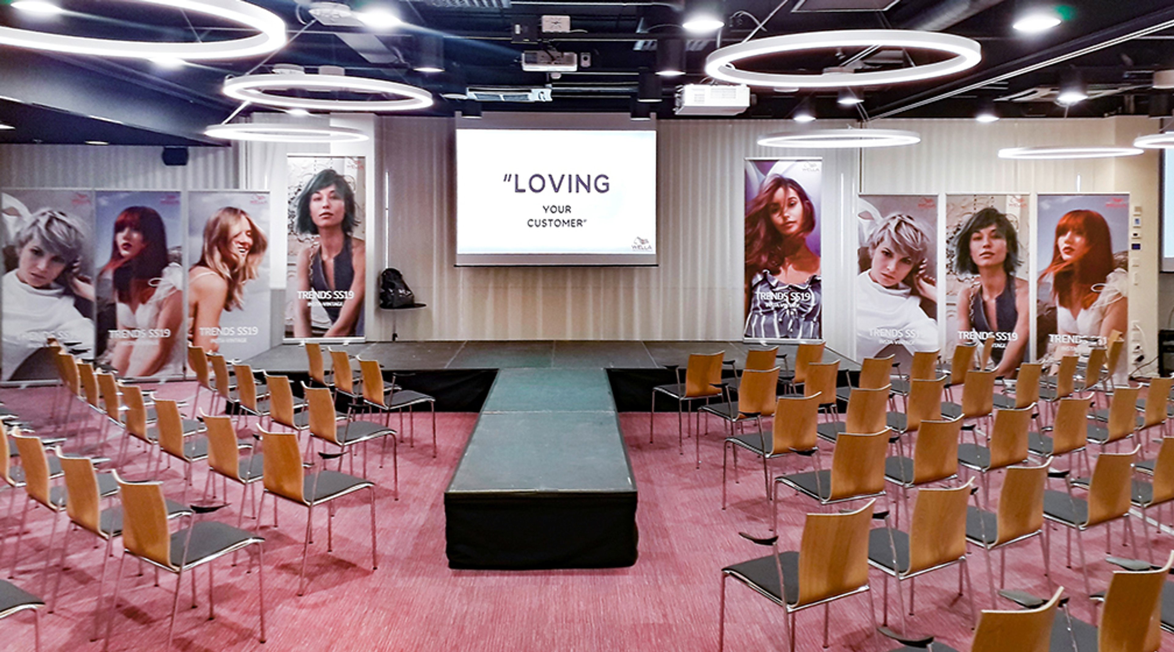 Empty conference room with wooden chairs, pink carpet, center aisle and black podium. Fashion portraits on walls and presentation slide on screen.