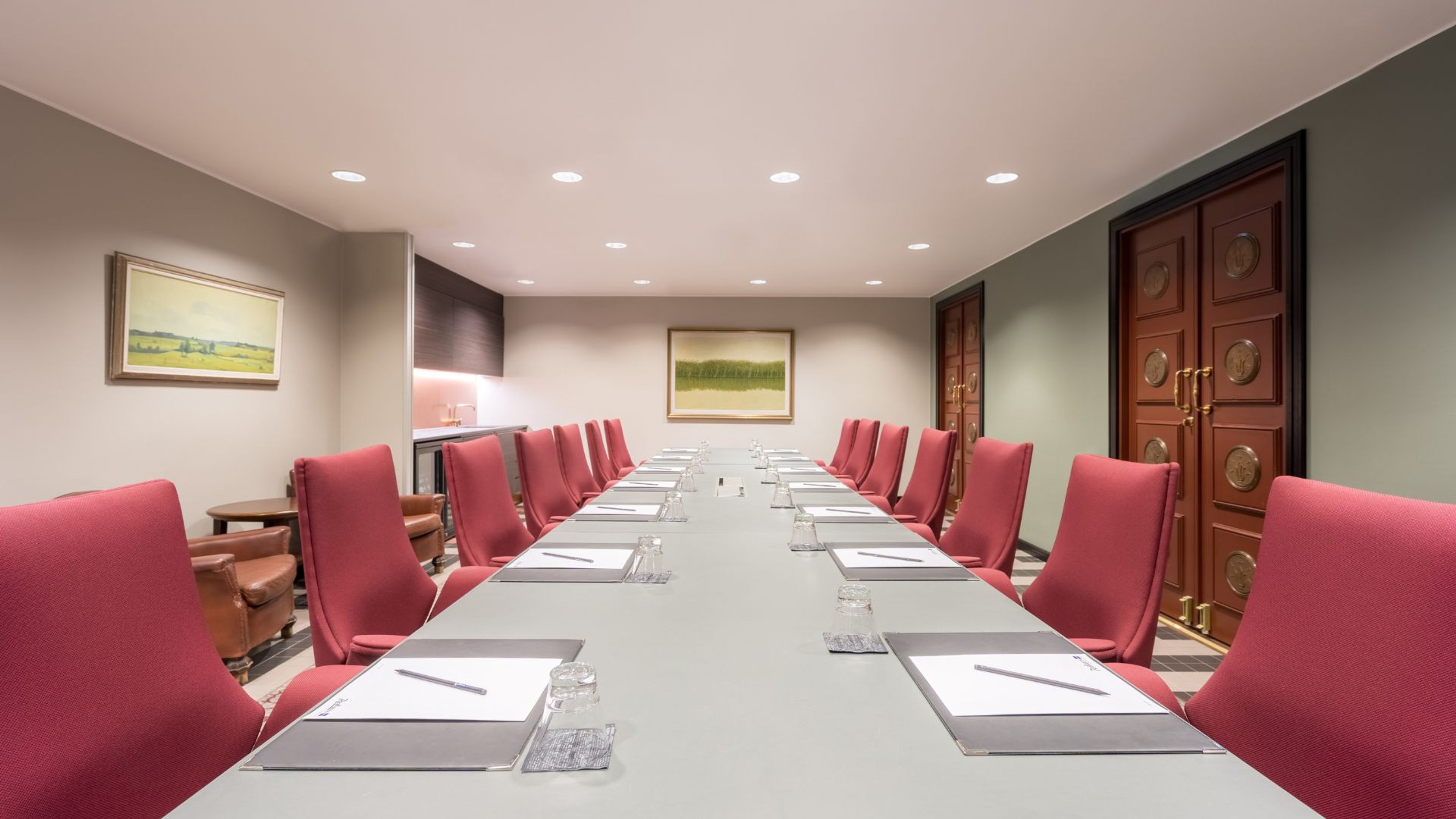 Kokoushuone, jossa on pitkä pöytä, punaisia tuoleja, puiset pariovet ja maisematauluja seinillä. Hyvin valaistu tila.