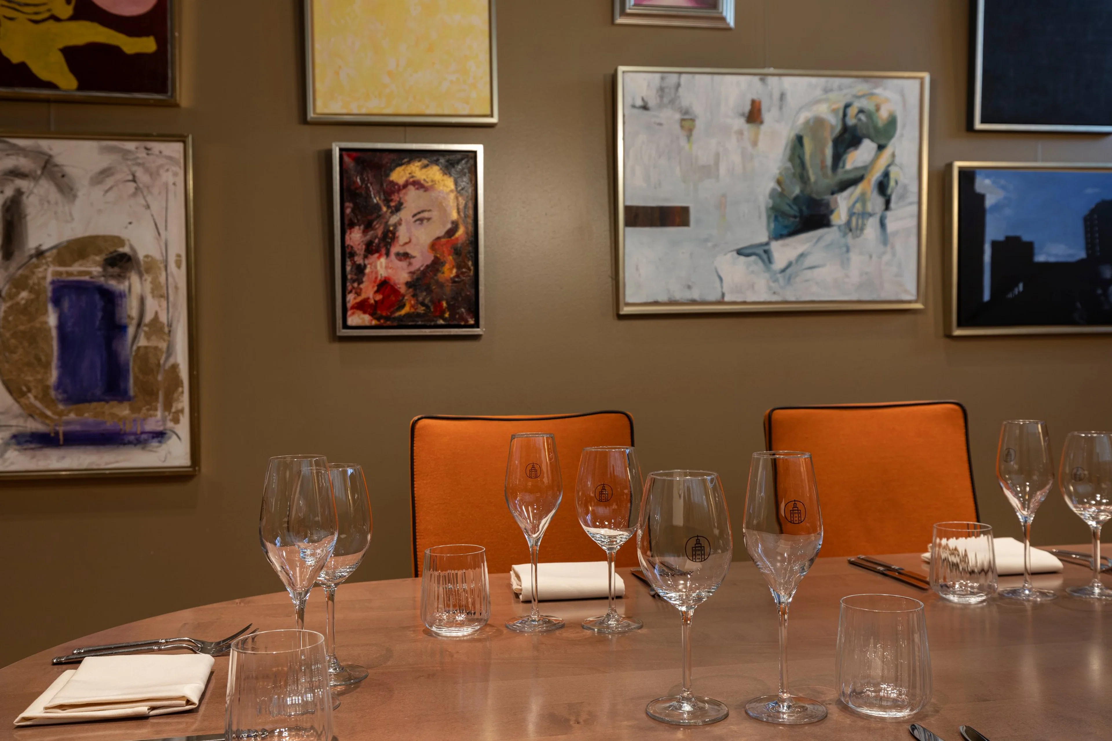 Ravintolan pöytä katettu laseilla, taustalla oranssit tuolit ja moderneja taideteoksia ruskealla seinällä.