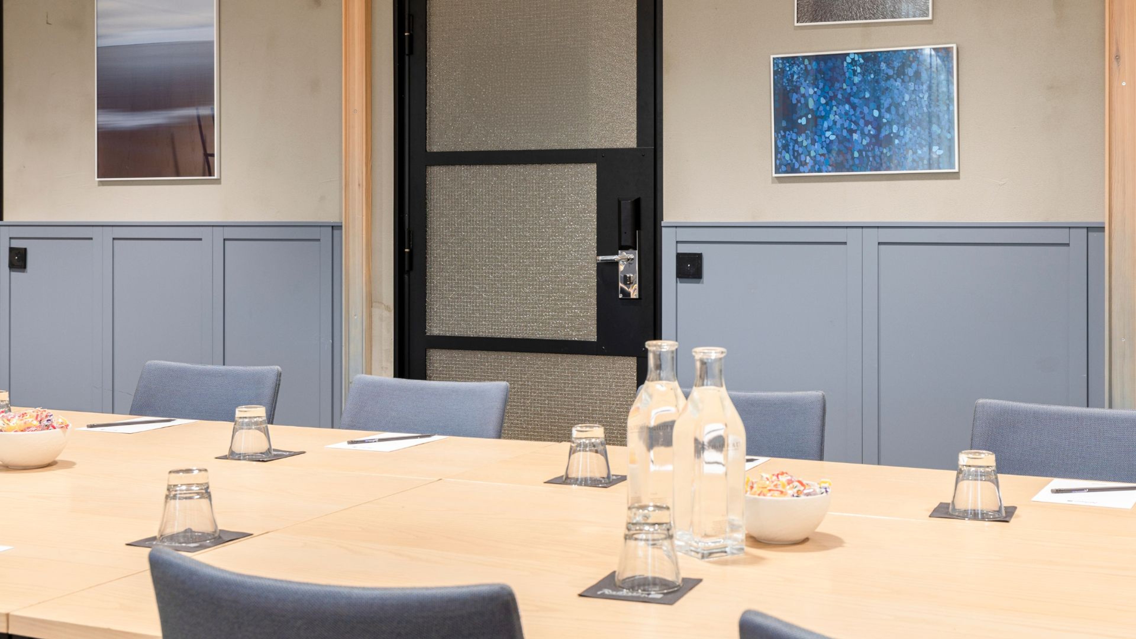 Neuvotteluhuone vaalealla puupöydällä, sinisillä tuoleilla, vesipulloilla ja karamellimaljoilla. Seinällä taideteoksia ja lasiovella.