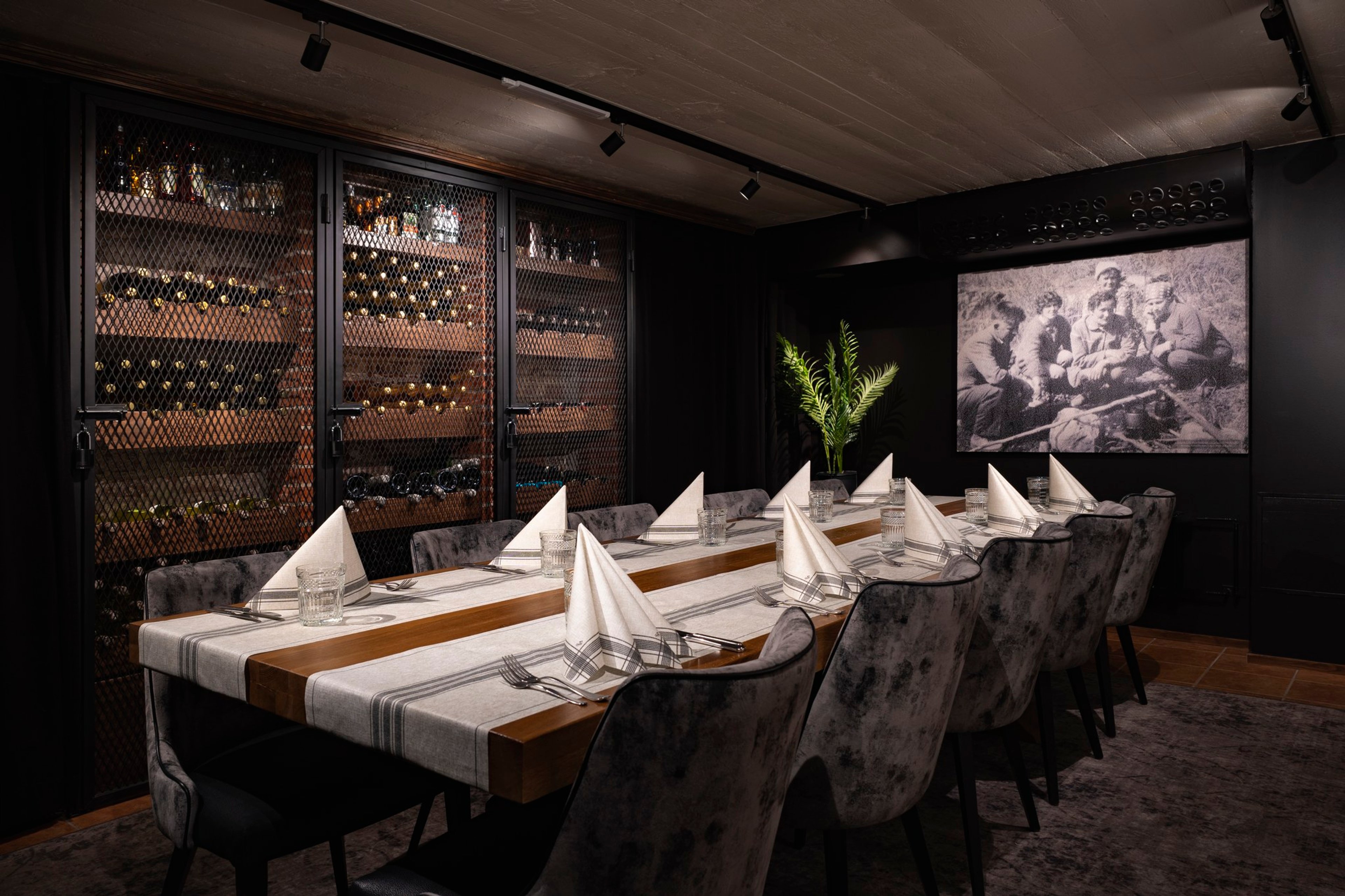 Tyylikäs yksityinen ruokailutila viinikaapilla, pitkä pöytä kattauksella, tummat samettiset tuolit ja historiallinen kuva seinällä.