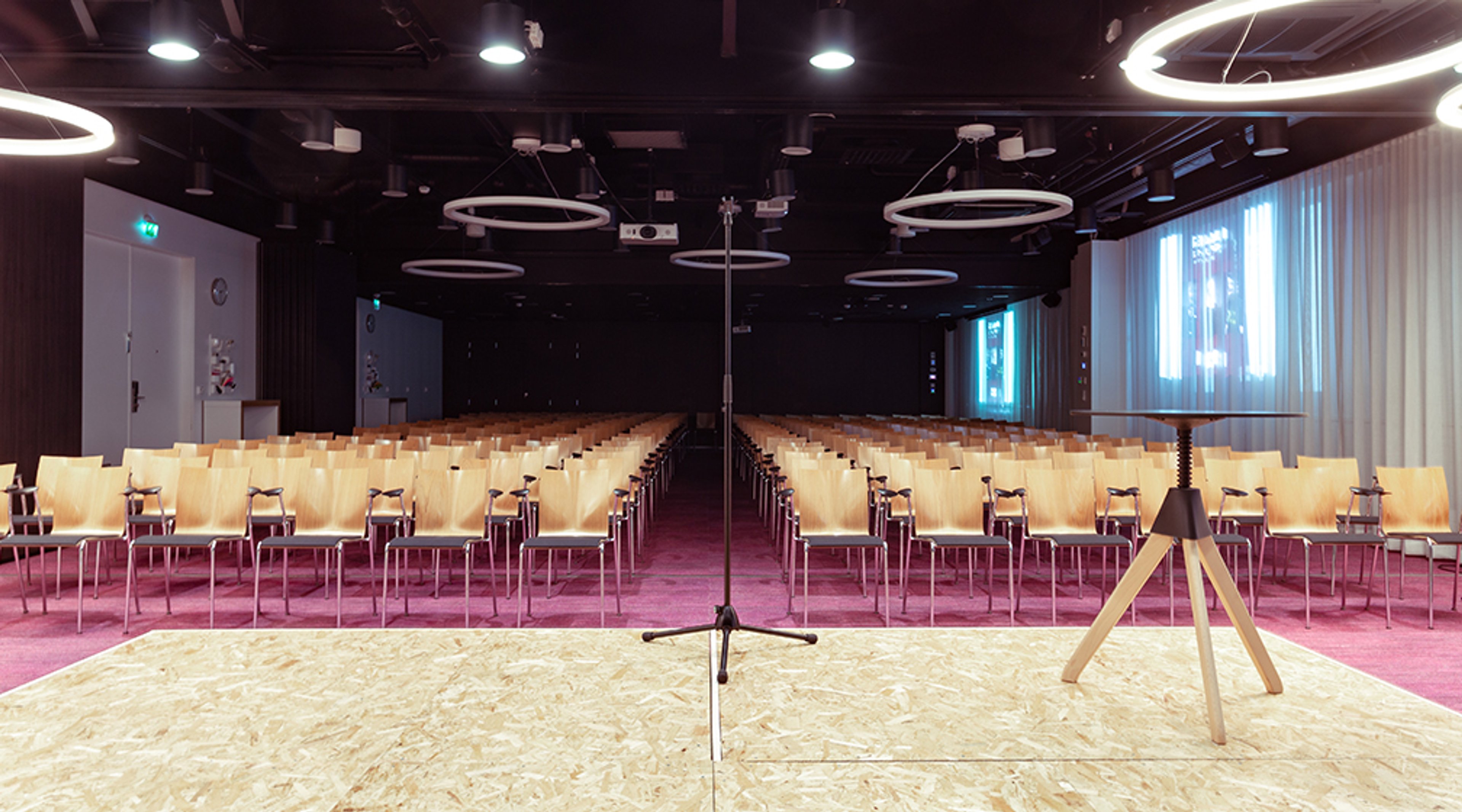 Modern auditorium with wooden chairs, pink carpet, and a wooden stage platform. Ceiling features ring-shaped lighting fixtures.