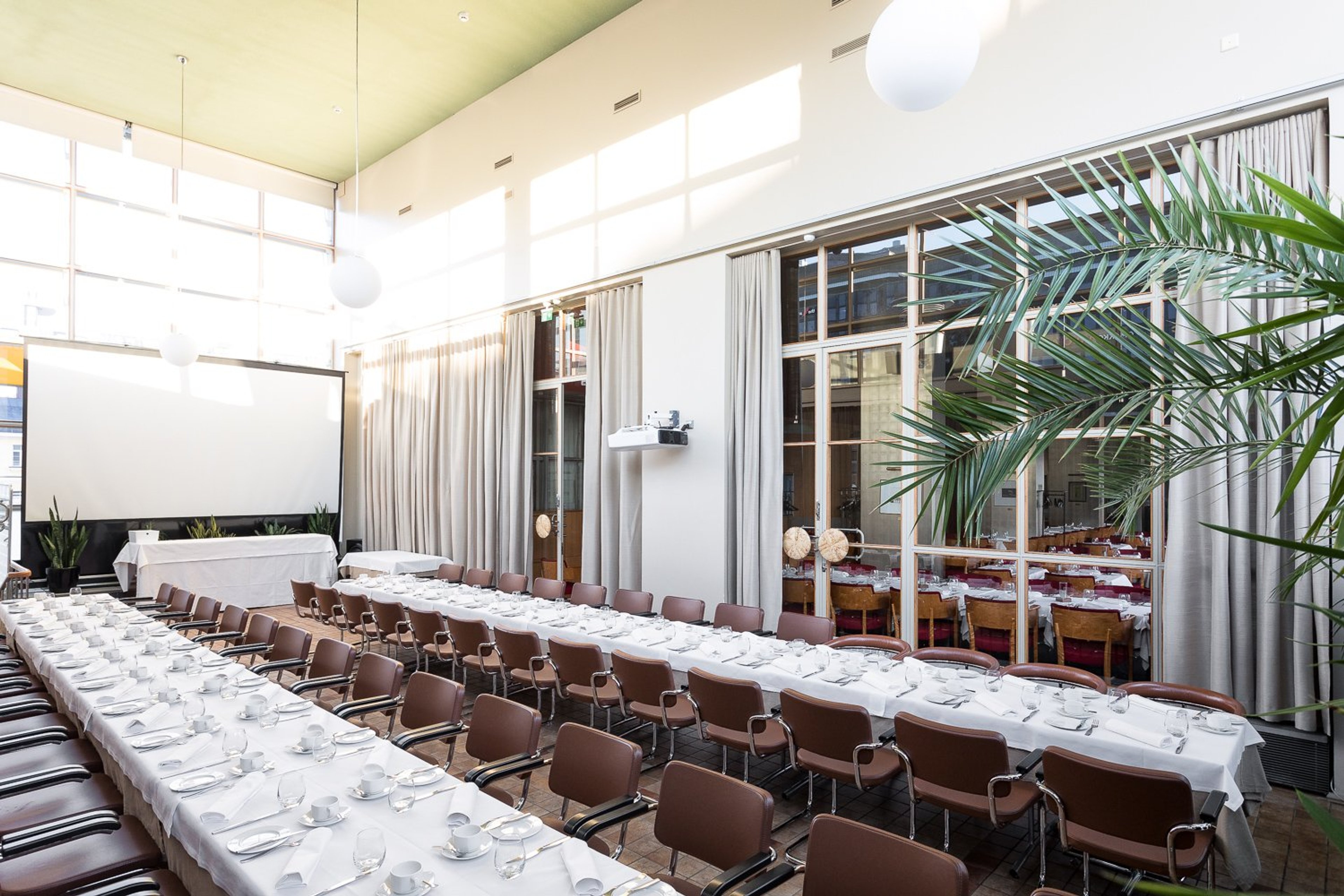 Ljus konferenslokal med långa bord täckta med vita dukar och kuvert. Bruna stolar, projektorduk och stora fönster. En grön palm i hörnet.
