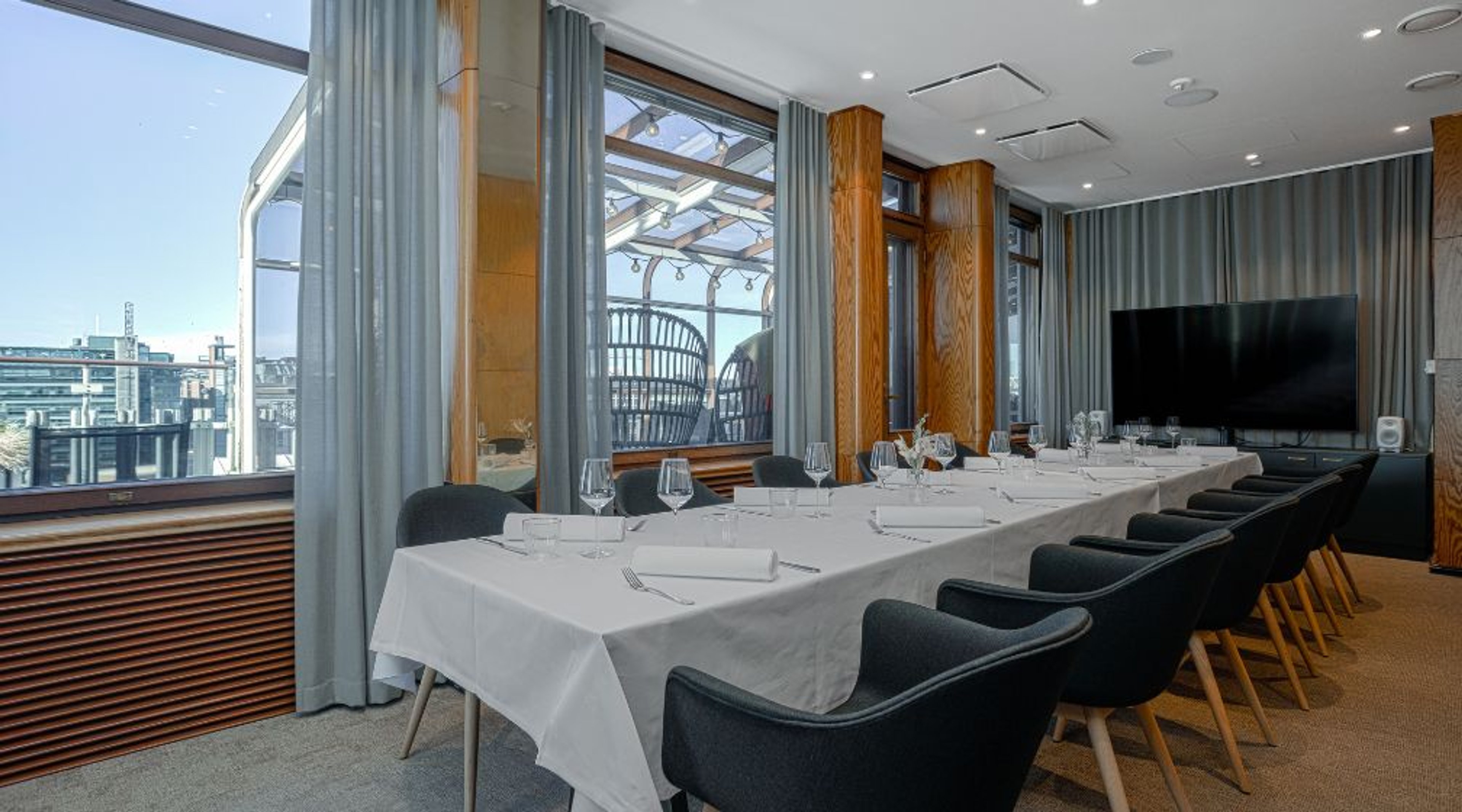 Ljust mötesrum med ett långt bord med vit duk och grå stolar. Stora fönster med stadsutsikt. Rummet har en TV-skärm och träpelare.