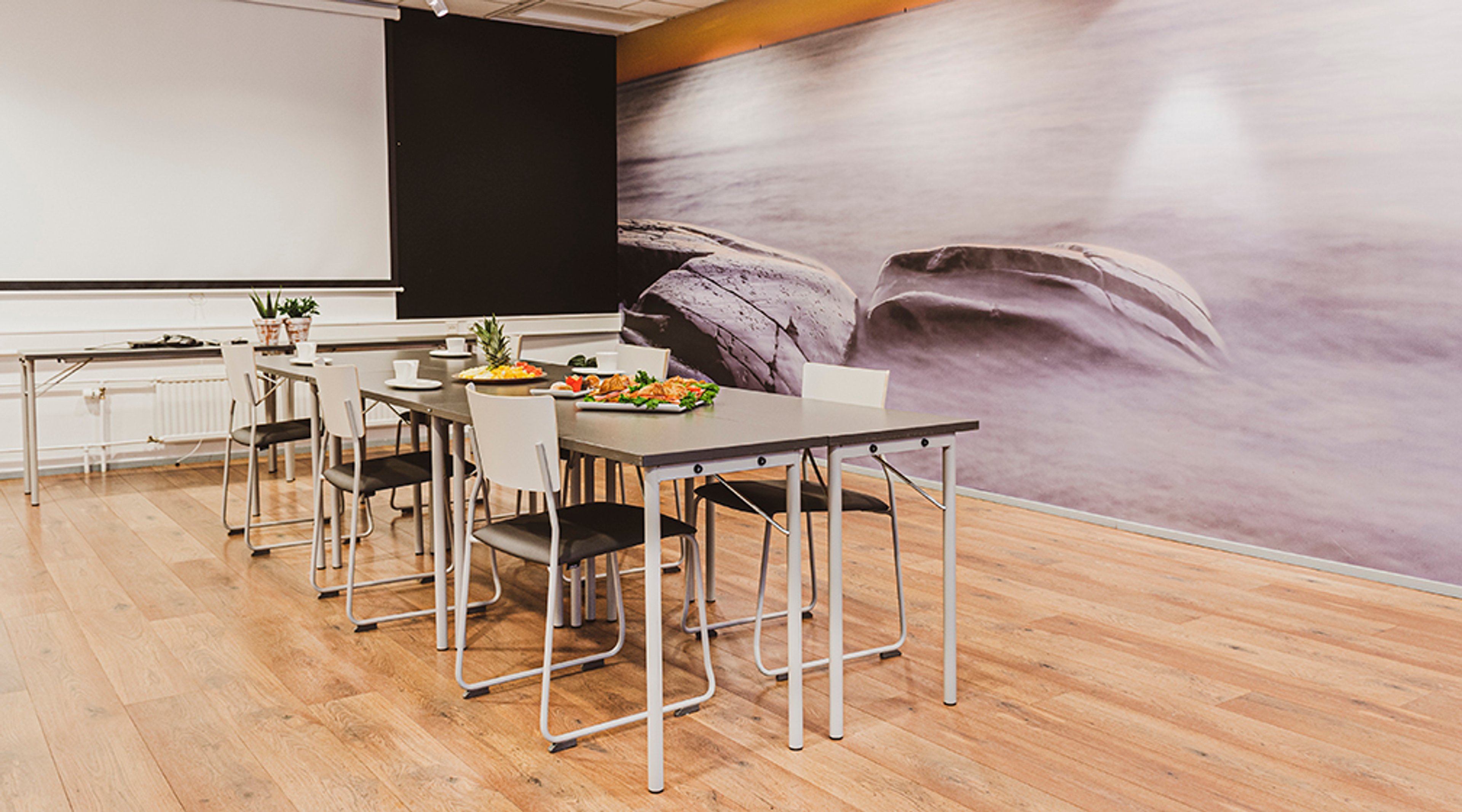 Kokoustila vaalealla puulattialla, jossa harmaat pöydät ja välipalat. Seinällä suuri kivikuvioinen maisemakuva ja valkokangas.