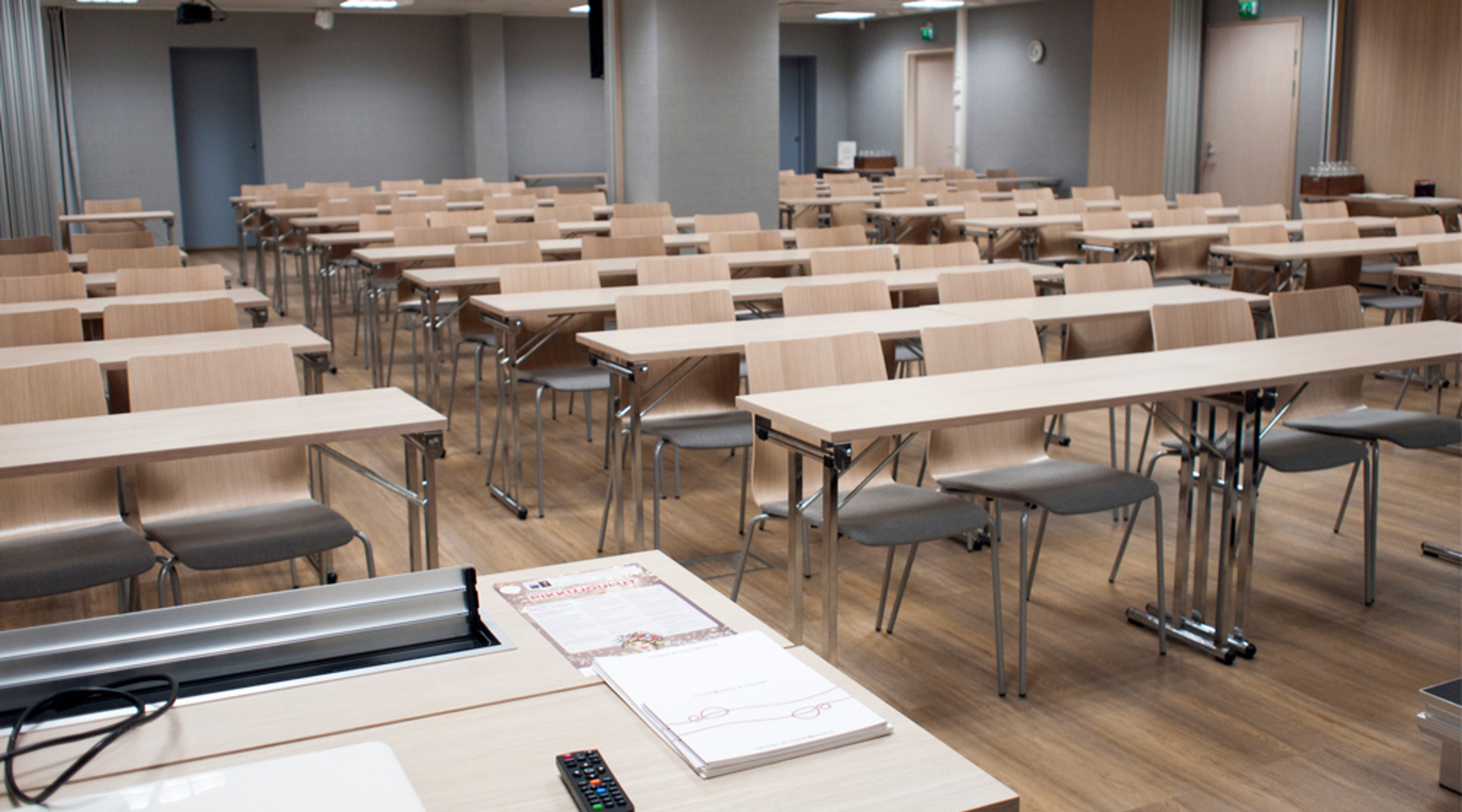 Luokkahuone tai kokoustila, jossa on rivejä vaaleita puisia pöytiä ja harmaita tuoleja. Edessä on opettajan pöytä, jolla on papereita ja kaukosäädin.