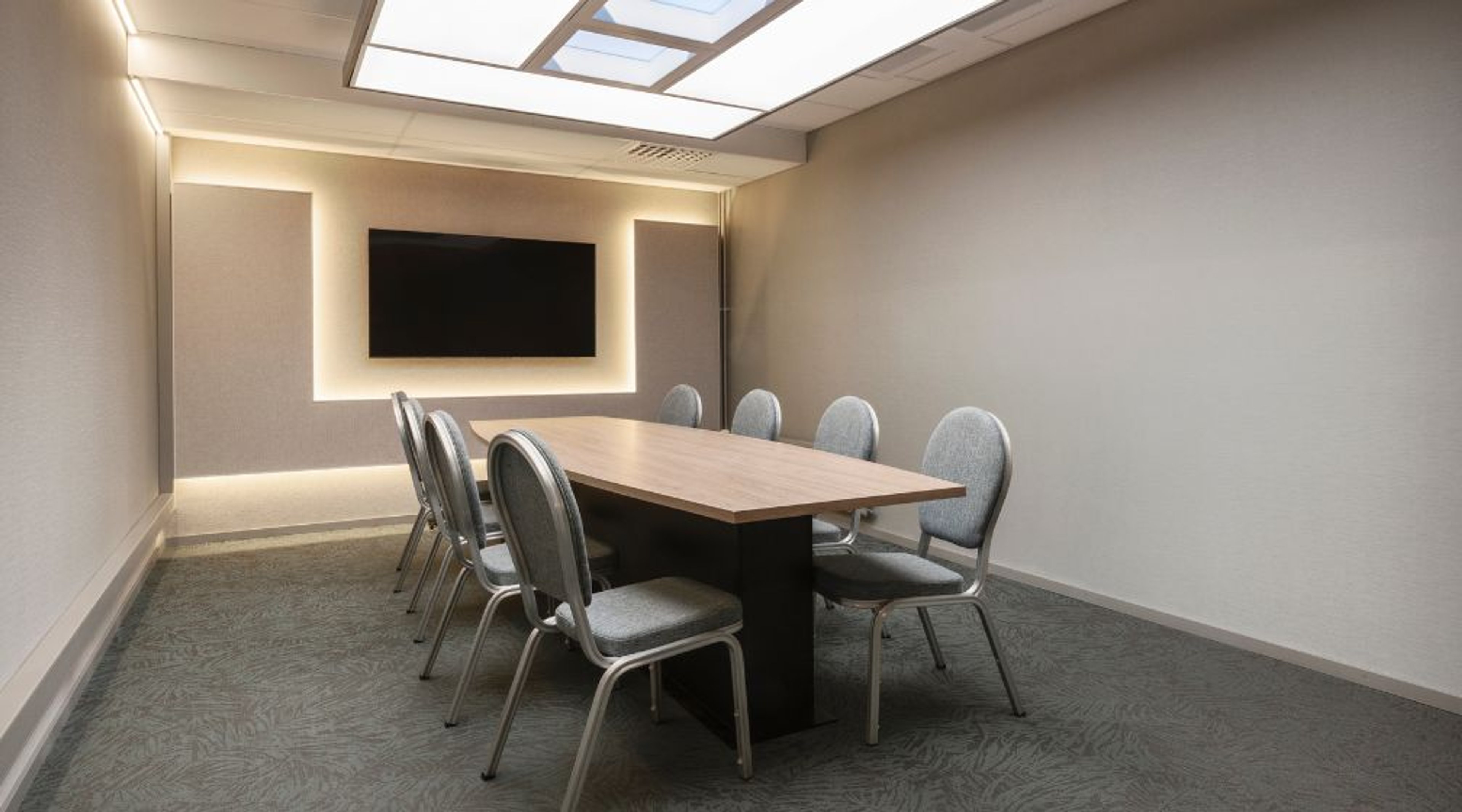 Moderni neuvotteluhuone puupöydällä, harmailla tuoleilla ja seinään upotetulla näytöllä. Huoneessa on hyvä valaistus ja tasainen lattia.
