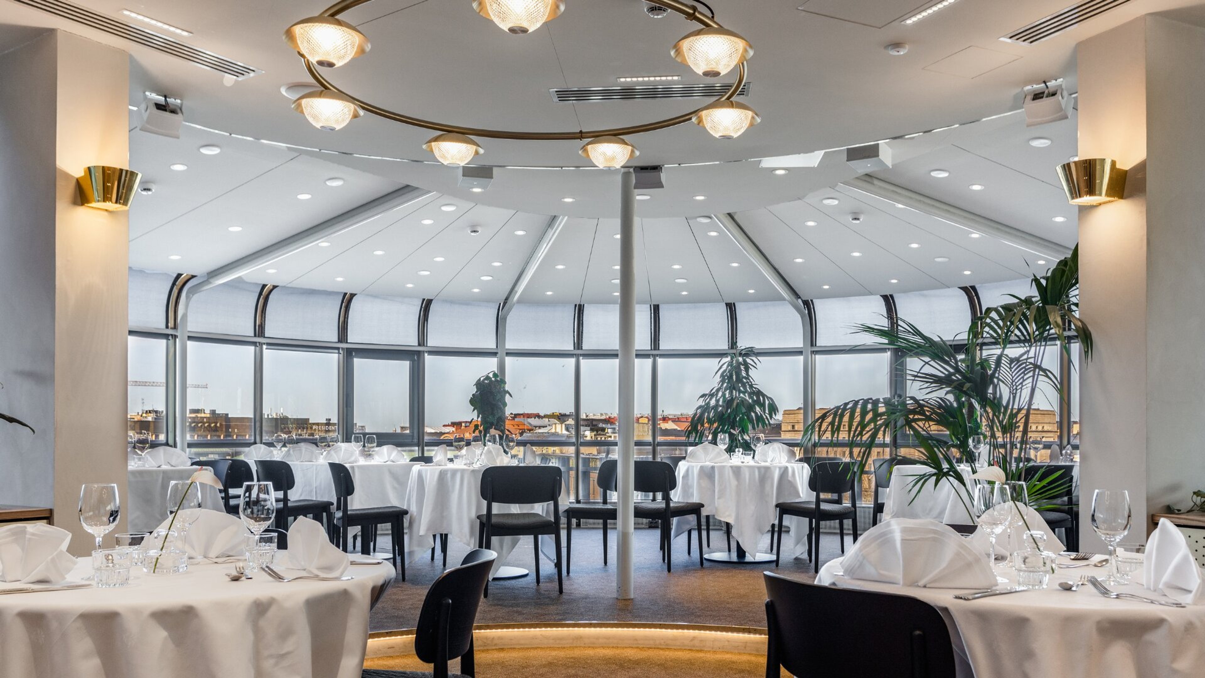 Tyylikäs pyöreä ravintolasali, jossa valkoiset pöydät ja mustat tuolit. Suurista ikkunoista näkyy kaupunkimaisema. Korkea katto, koristeellinen valaistus.