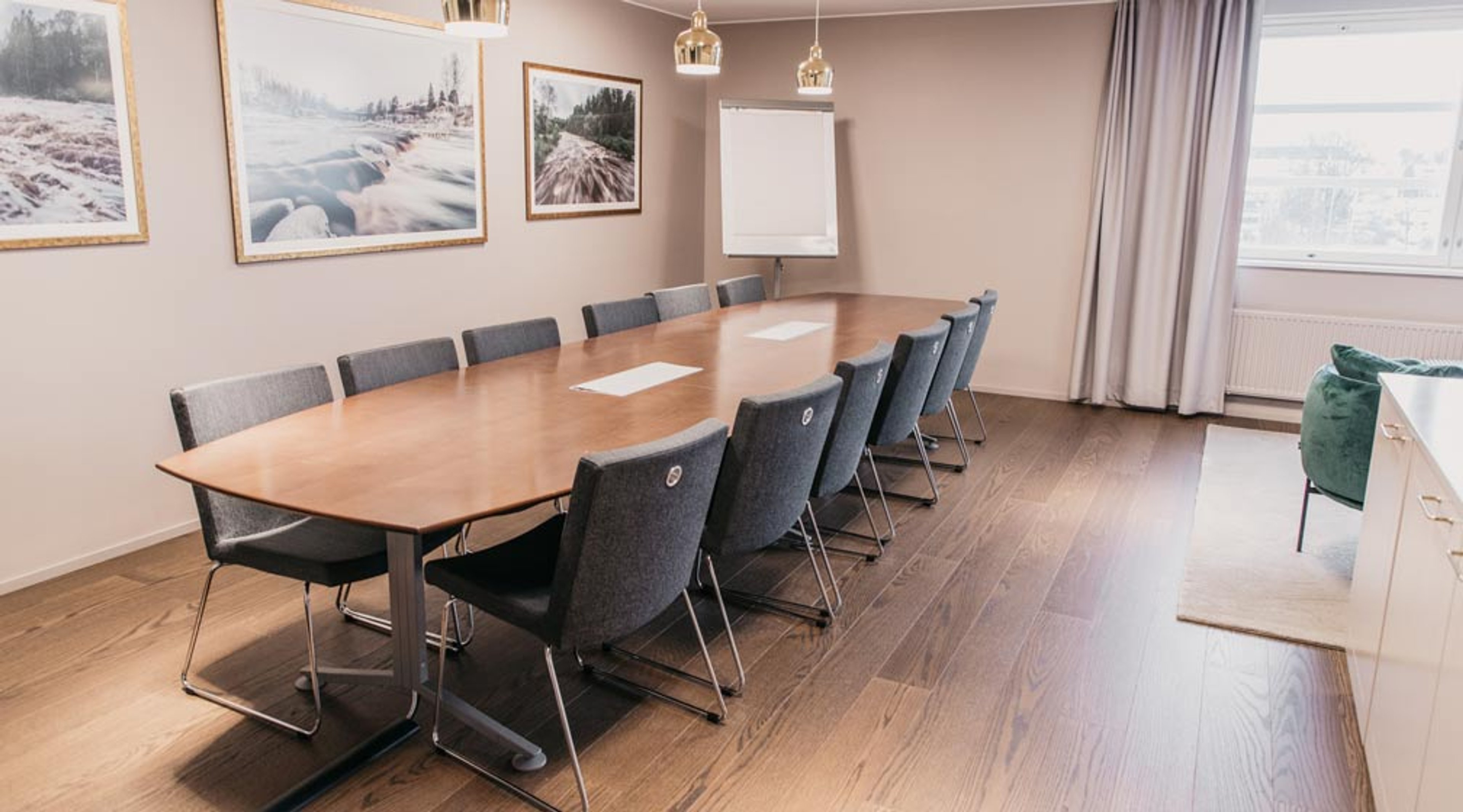 Kokoushuone, jossa pitkä puinen pöytä ja harmaat tuolit, seinillä luontokuvia, taululehtiö ja ikkuna. Kovapuulattia ja luonnonvalo.