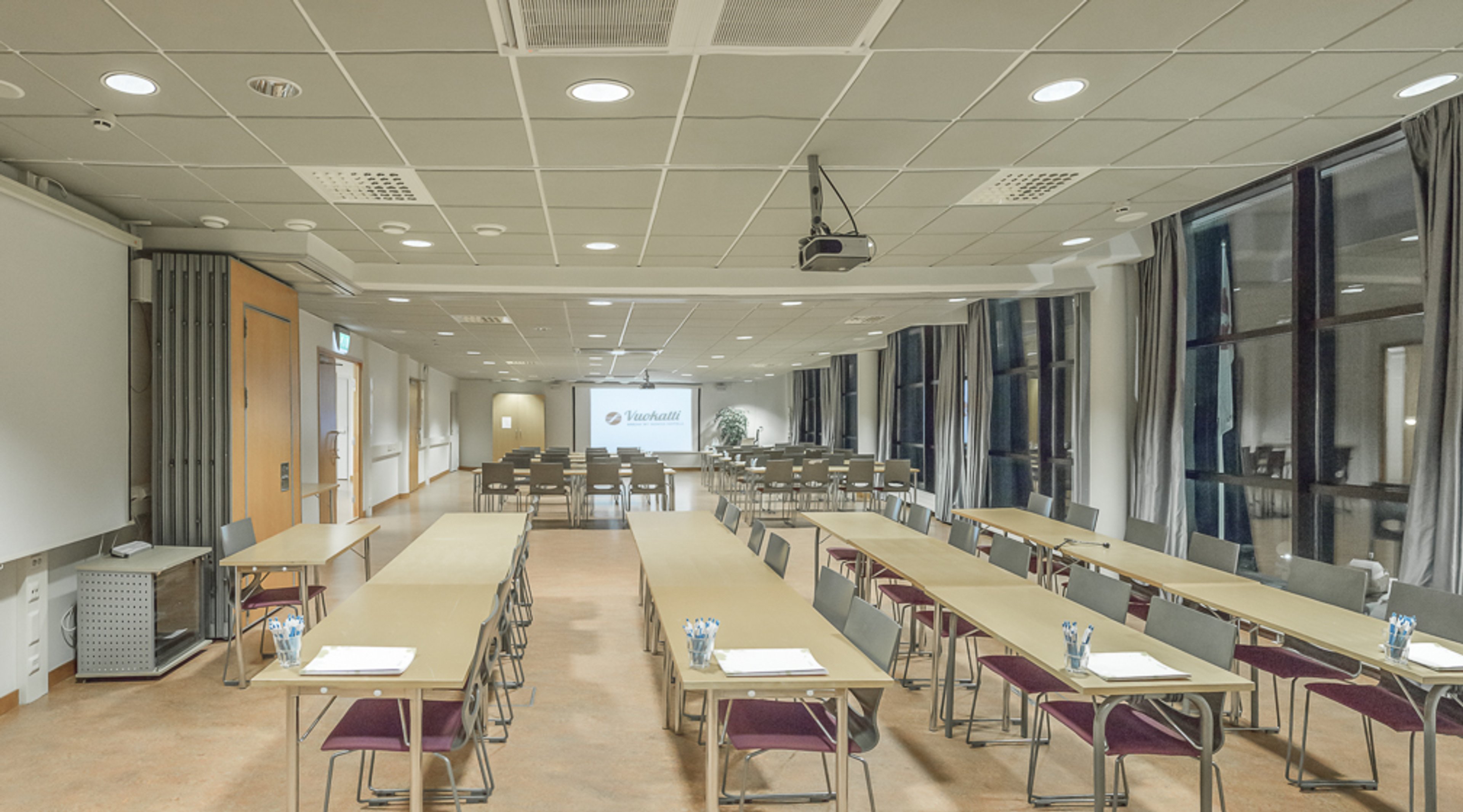 Valoisa kokoustila, jossa pitkiä pöytiä luokkamuodostelmassa, projektorikangas edessä ja suuret ikkunat sivuseinällä. Tila on avara ja tasalattiainen.