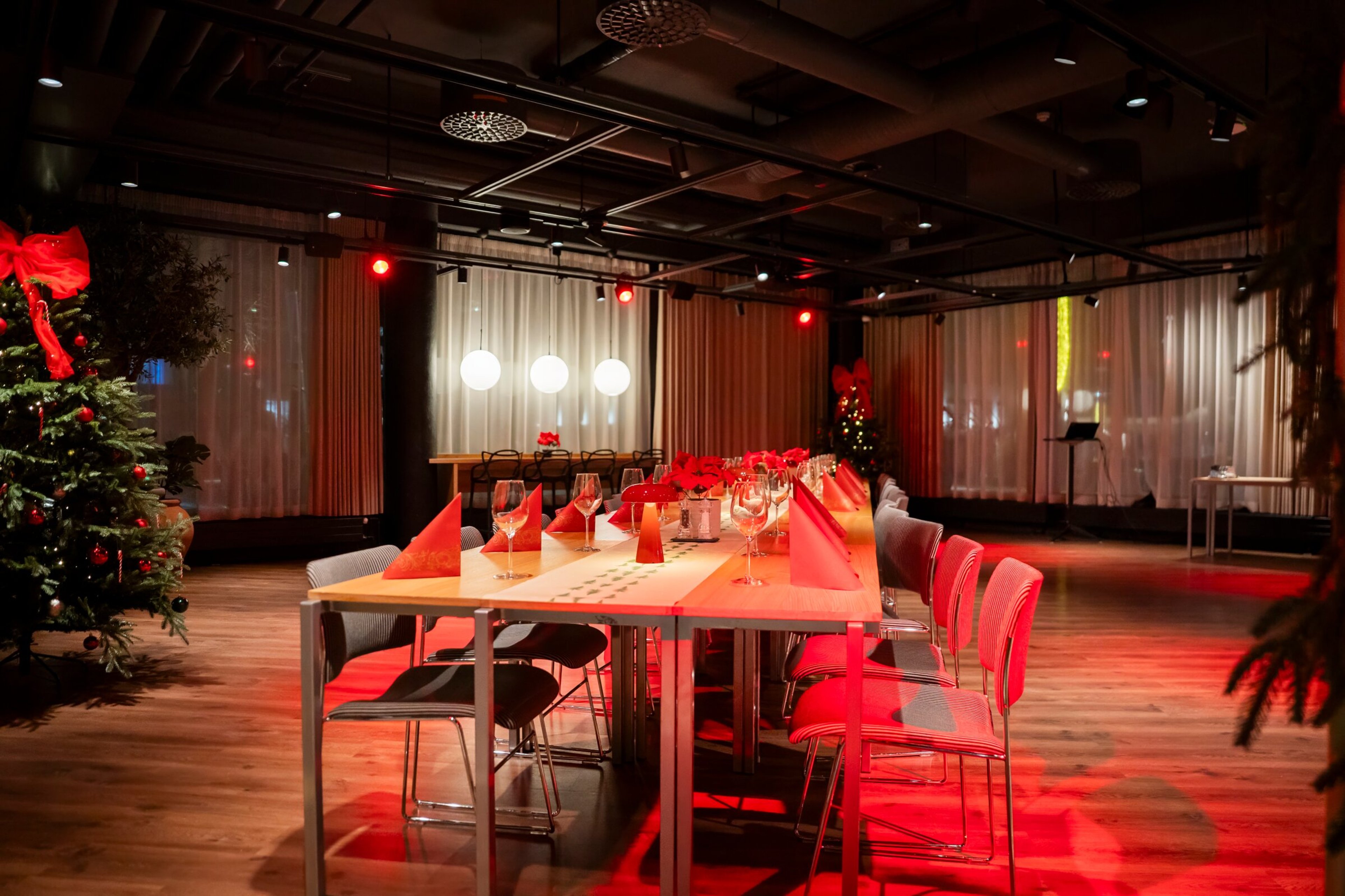 Juhlatila, jossa pöytä katettuna punaisilla lautasliinoilla, joulukuusi koristeluineen ja puulattia. Tilassa punaiset valot ja valkoisia verhoja.