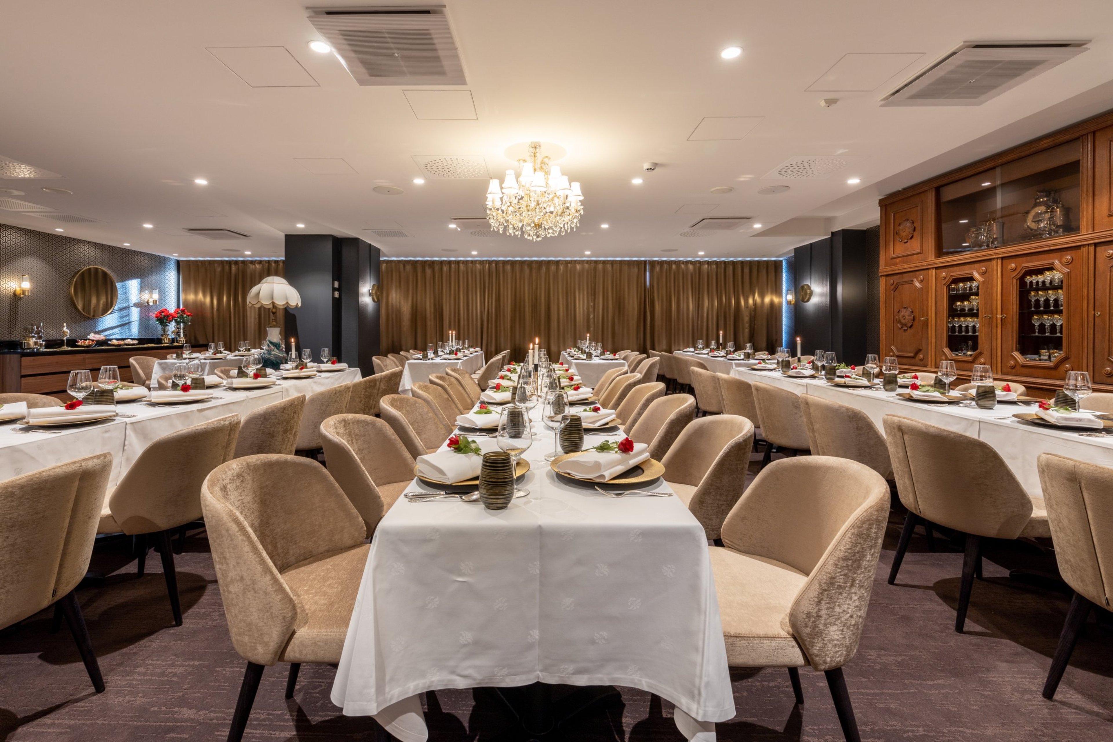 Tyylikäs ravintolasali, jossa valkoliinaiset pöydät, beiget tuolit, kristallikruunu katossa ja puinen kaappi sisustuksessa.