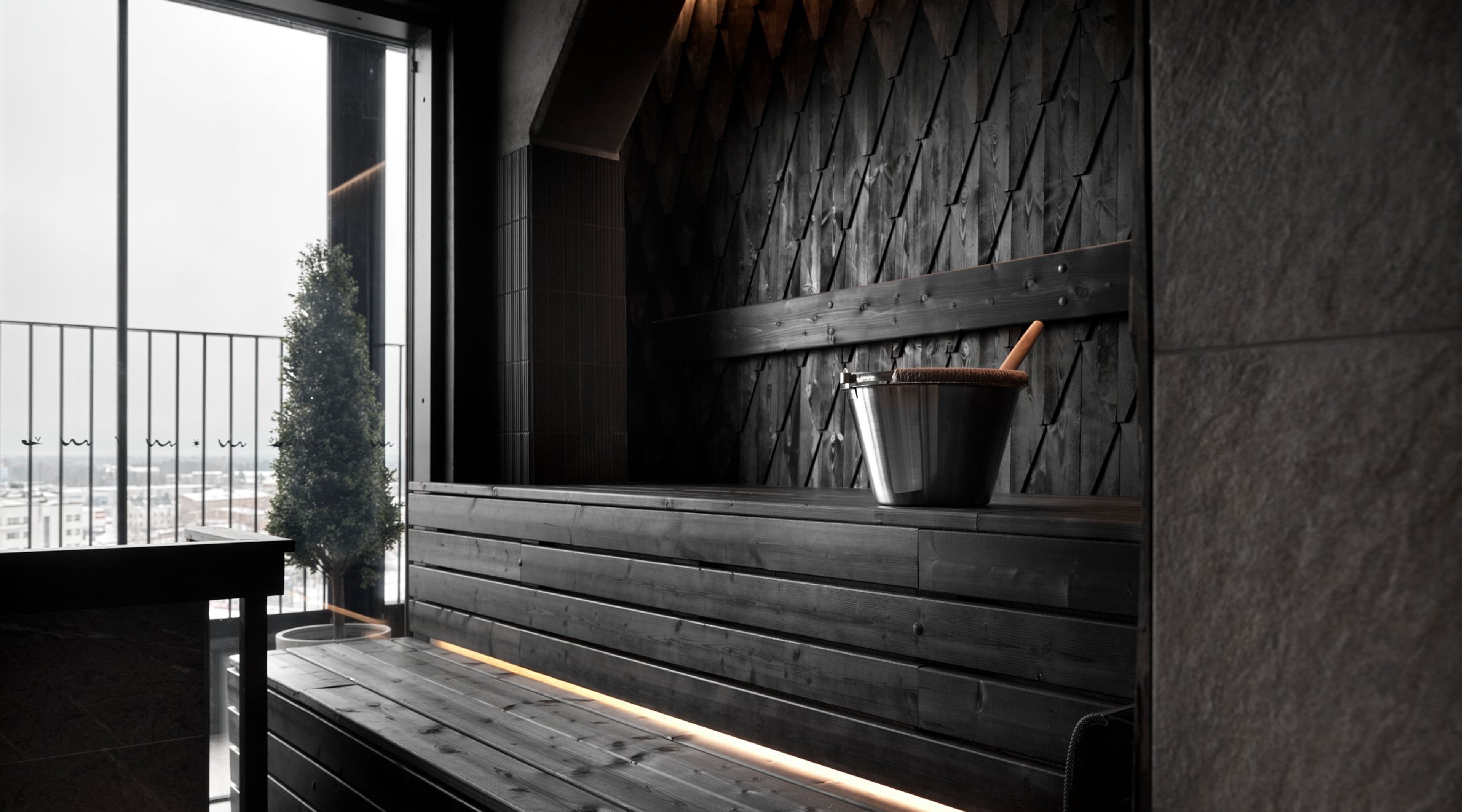 Moderni tumma sauna, jossa puupenkit ja metallinen kiulu. Suuresta ikkunasta avautuu näkymä kaupungin ylle.